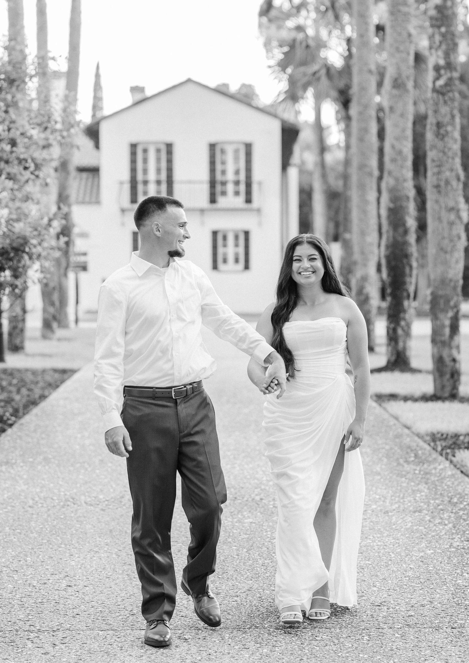 Black and white photo of a couple holding hands walking down a tree-lined path toward an elegant white estate.