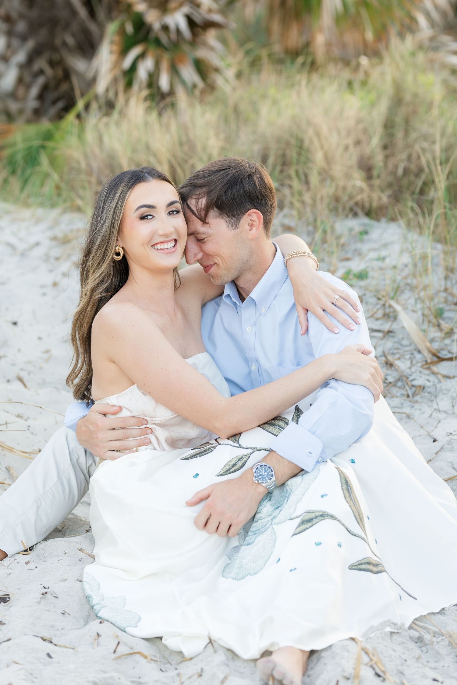 Romantic couple embracing on sandy beach, woman in floral dress smiling, man wearing blue watch.