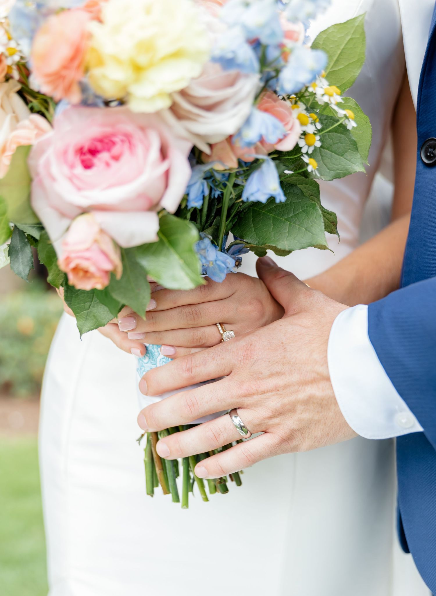 Close-up view of wedding bouquets with pink roses and hands showing engagement rings against blue attire.