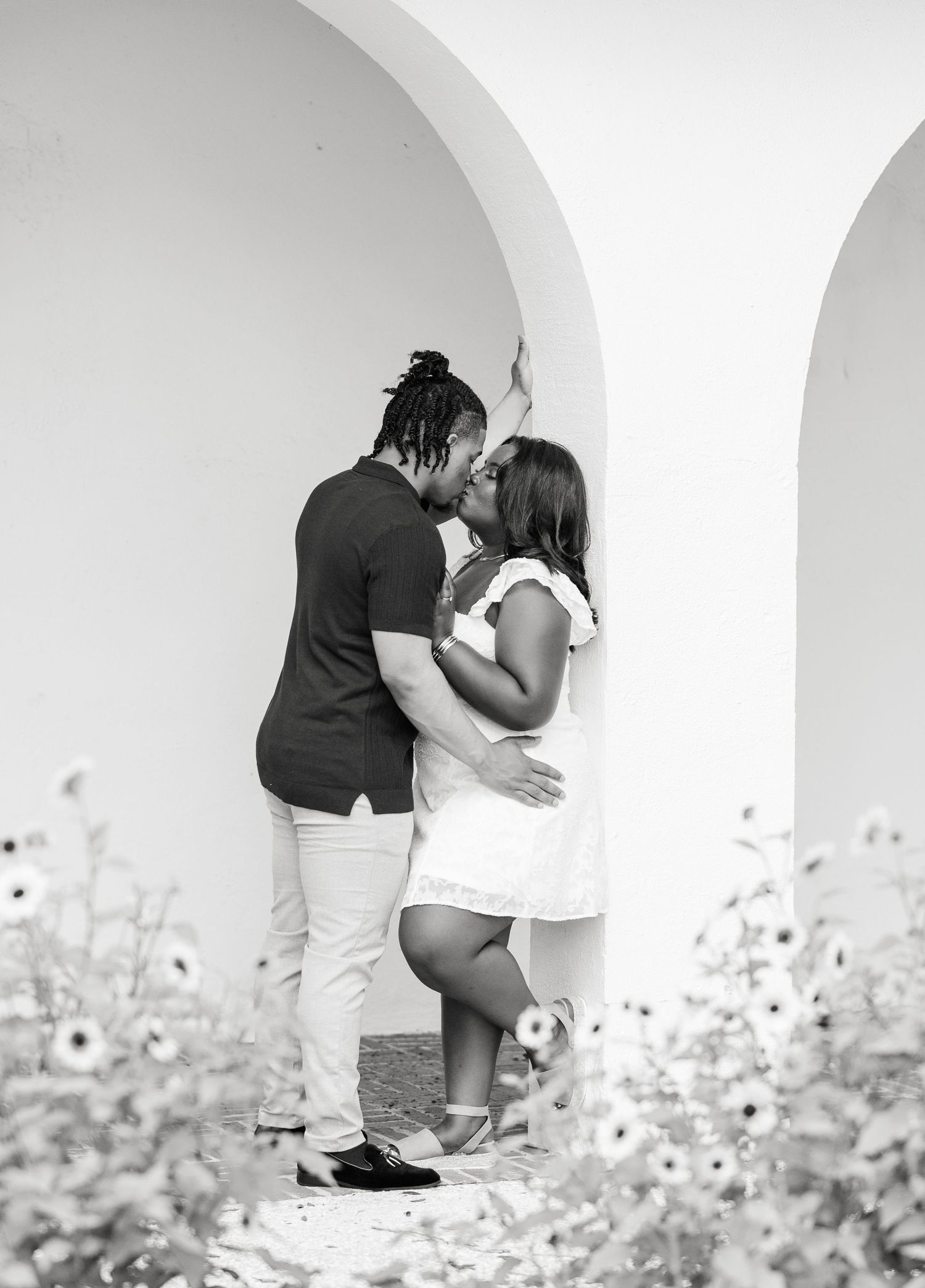 Black and white photo of a couple sharing a kiss under white arched columns with flowers.