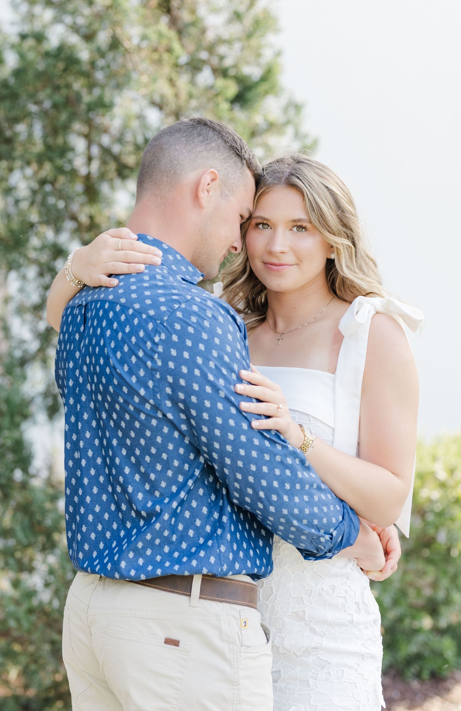 Couple sharing a tender moment, woman in white dress embracing man in blue patterned shirt outdoors.