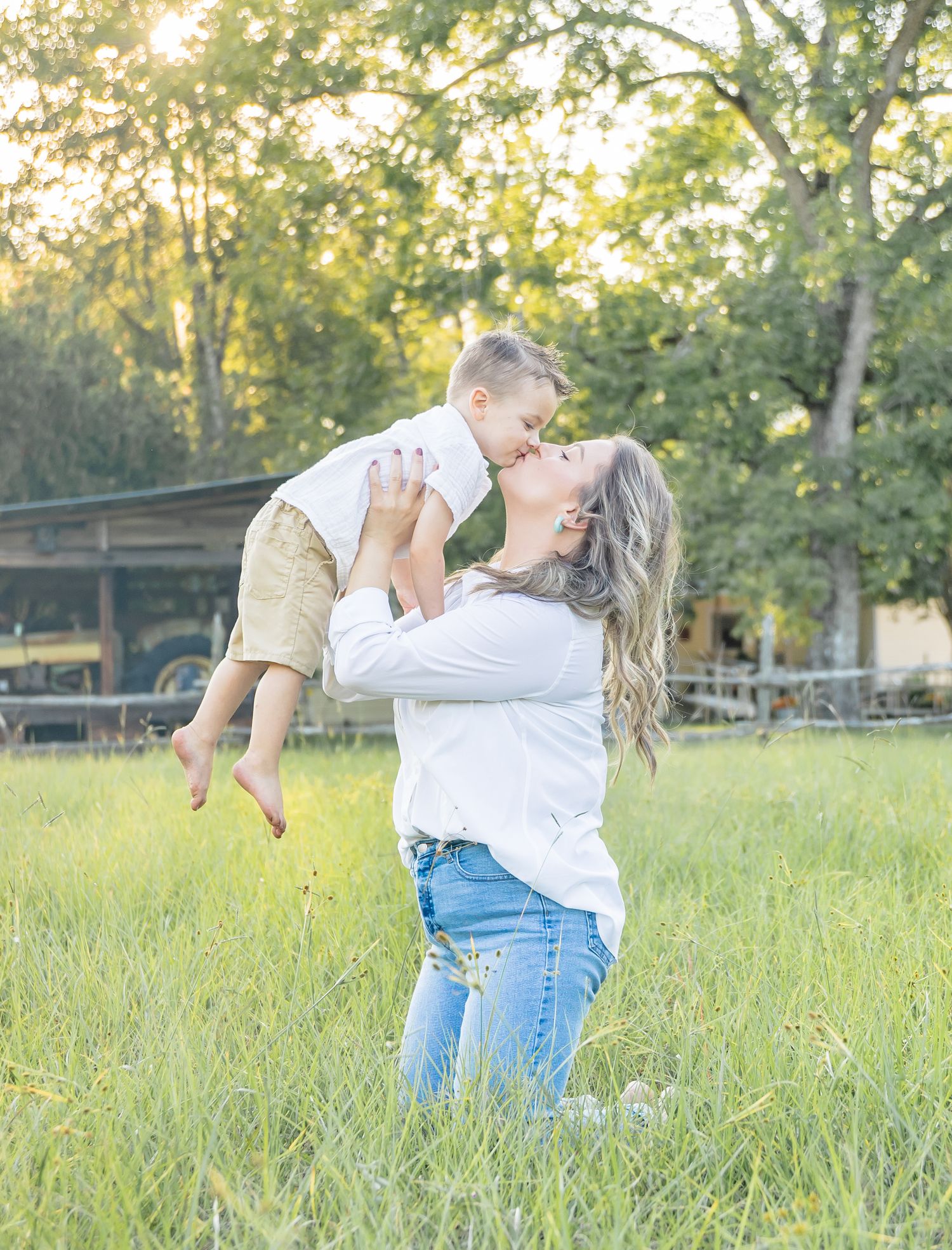 A couple dances playfully in a sun-drenched meadow with tall grass and trees in the background during golden hour.