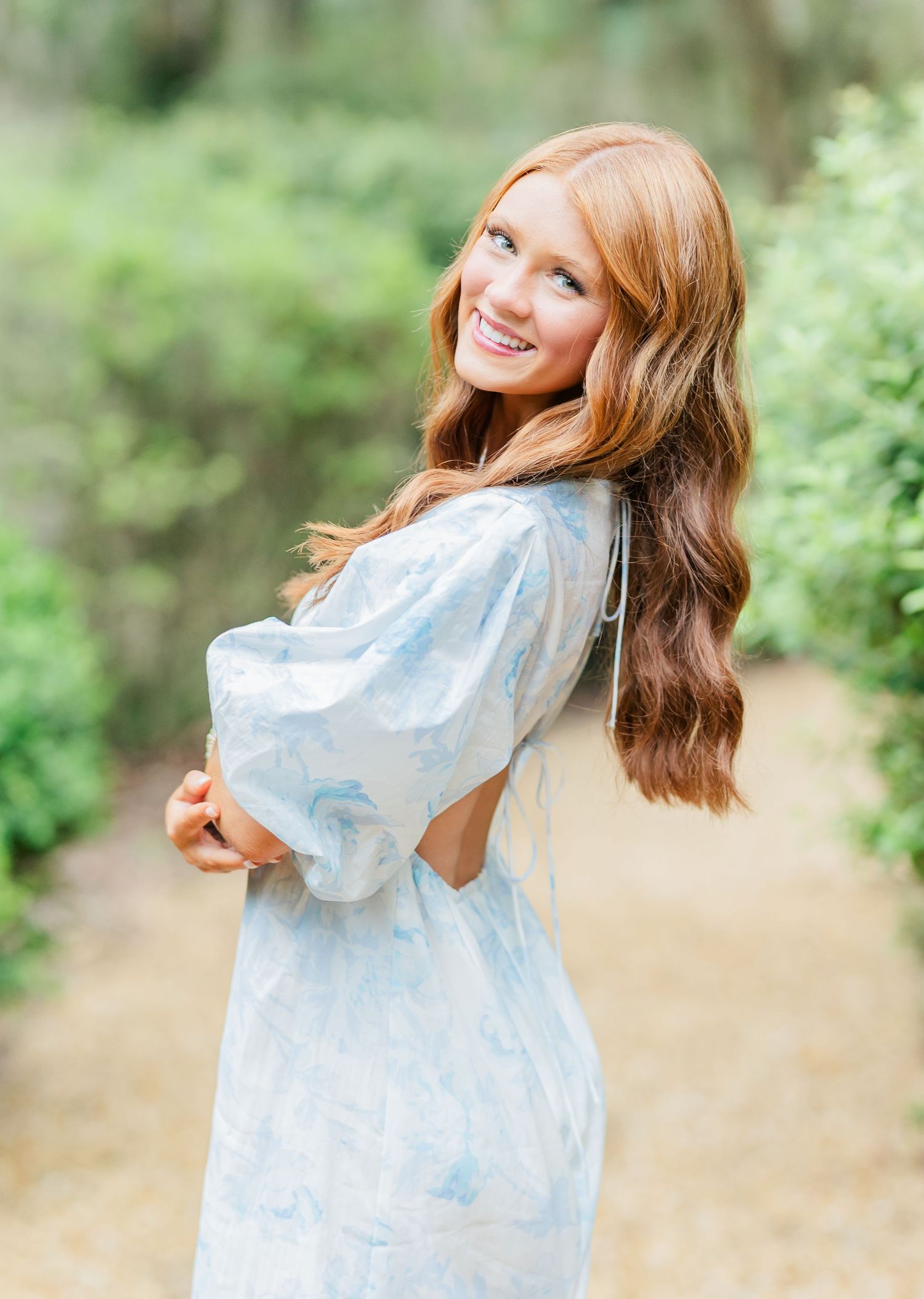 A figure in a light blue dress poses playfully against a soft natural background with bokeh effect.