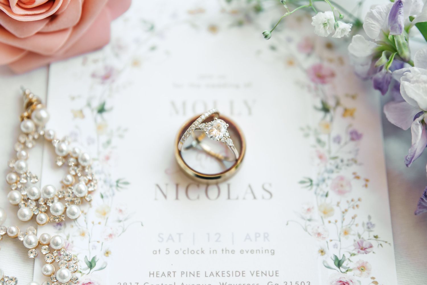Wedding rings and bridal jewelry arranged on floral invitation card with pink and purple flowers.