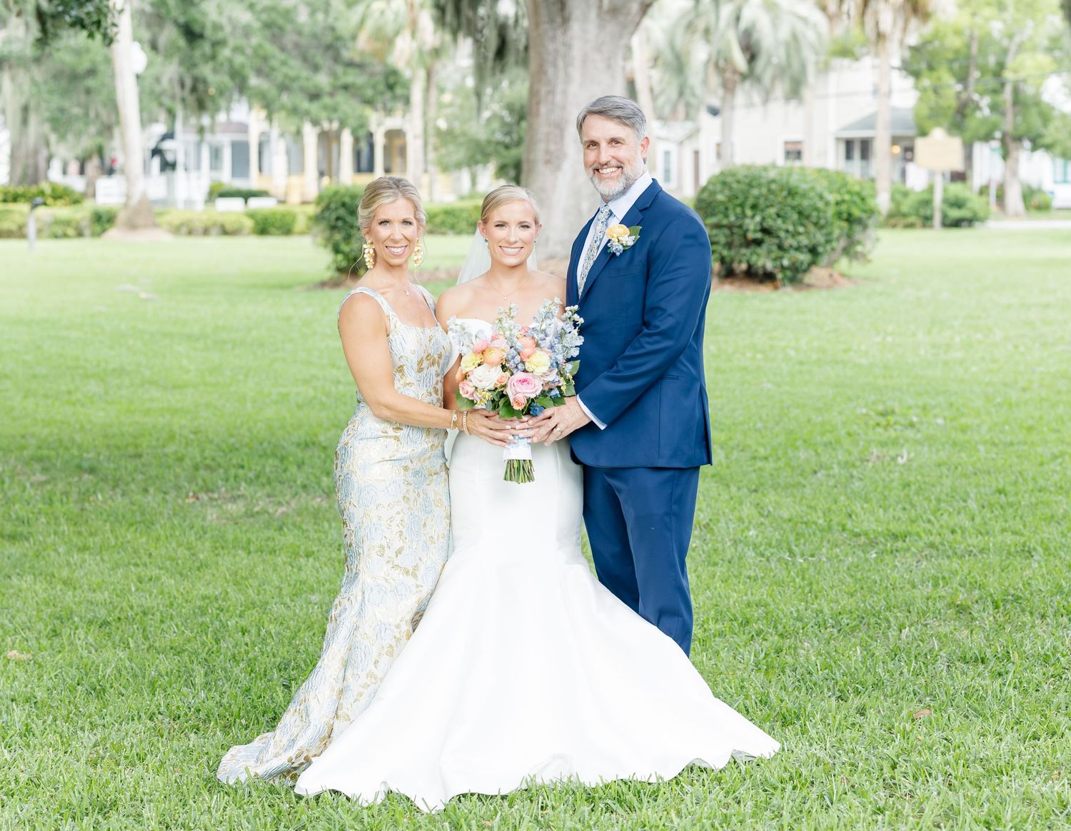 Three people in formal wedding attire pose together on a green lawn with houses and trees visible in the background.