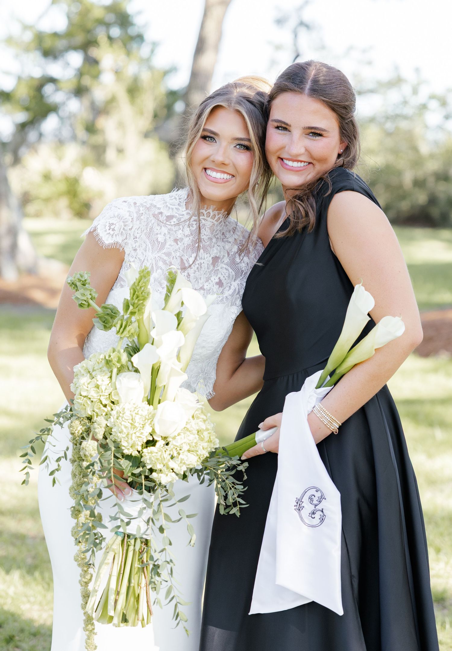 Bride in white lace gown holds floral bouquet beside bridesmaid in black dress holding monogrammed cloth outdoors.