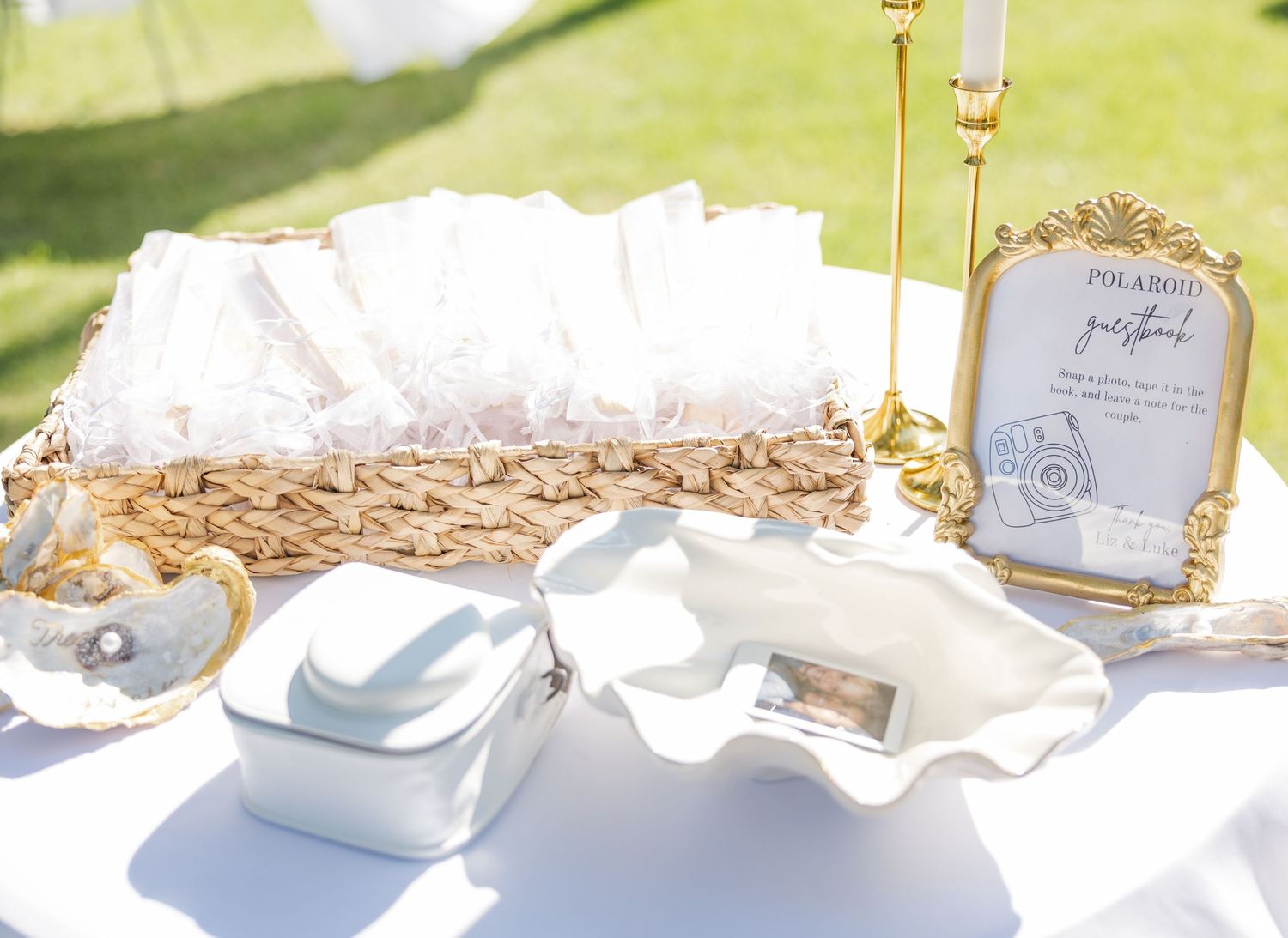 Elegant wedding Polaroid guestbook table with wicker basket, gold candlestick, framed sign, and white ceramic dishes outdoors.