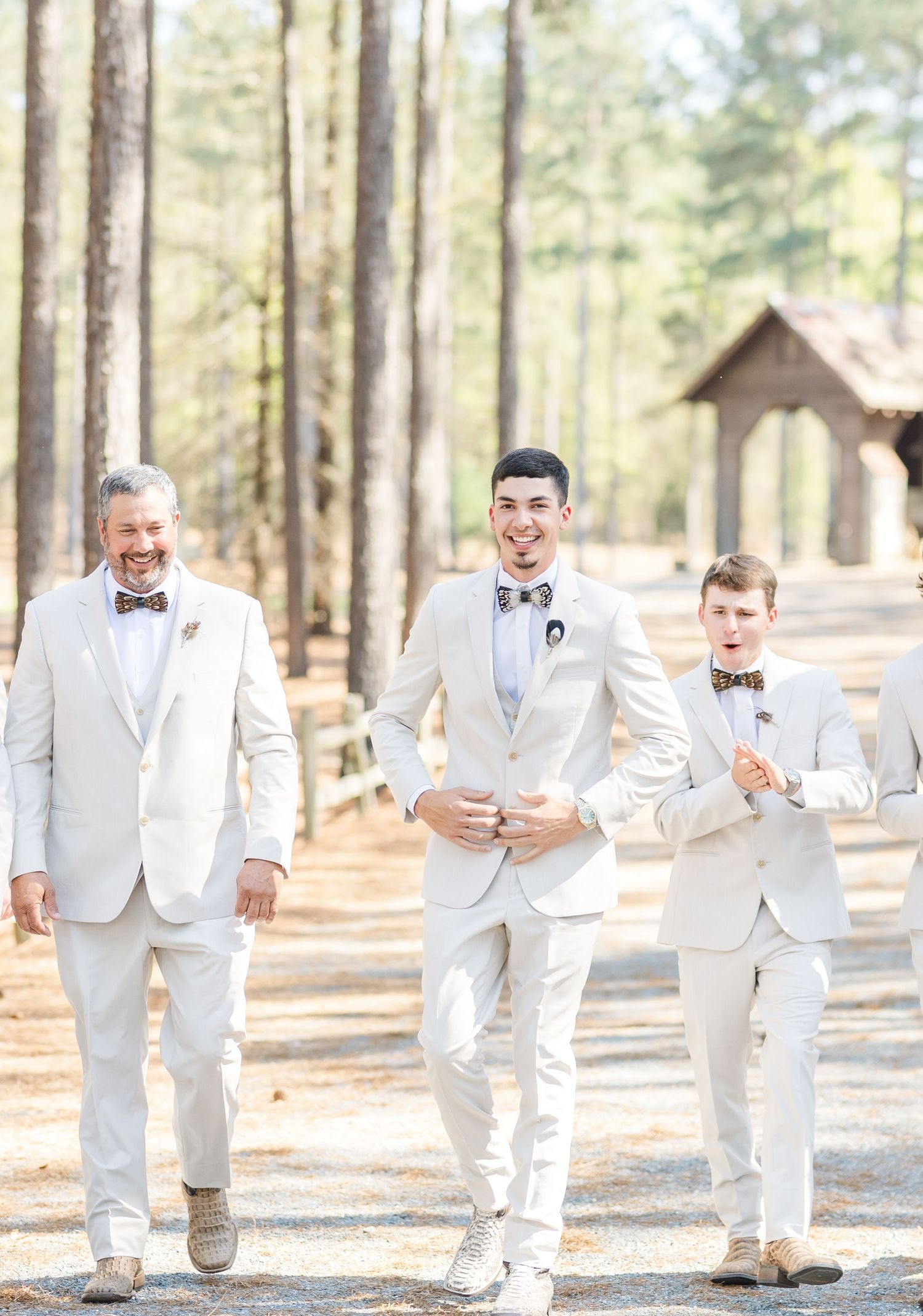 Groom and two groomsmen in ivory suits walk confidently along a sunlit pine forest path.
