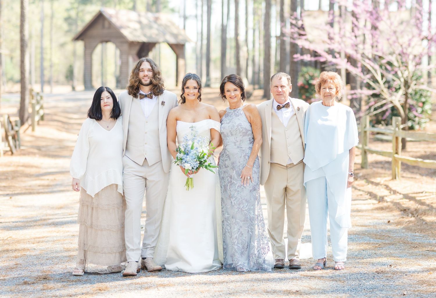 Wedding party of six poses together in neutral tones along a pine tree-lined venue path.