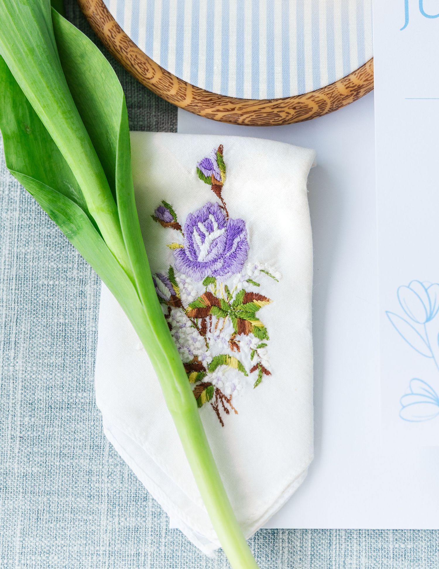 White embroidered napkin with purple floral design beside a green tulip stem on a light blue linen tablescape.