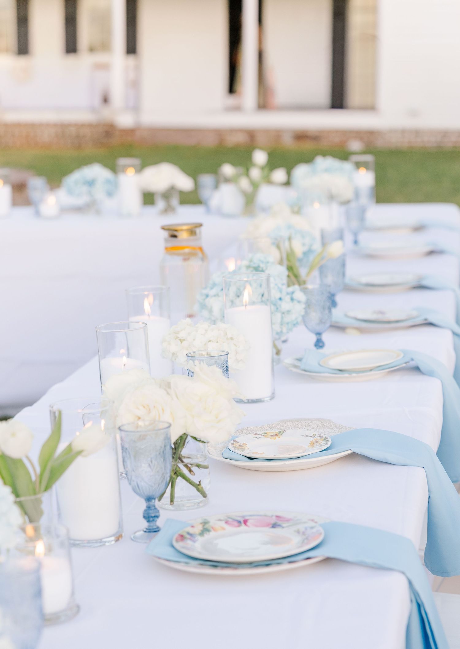 Elegant outdoor wedding reception table with white linens, blue hydrangeas, white roses, candles, and vintage floral china.