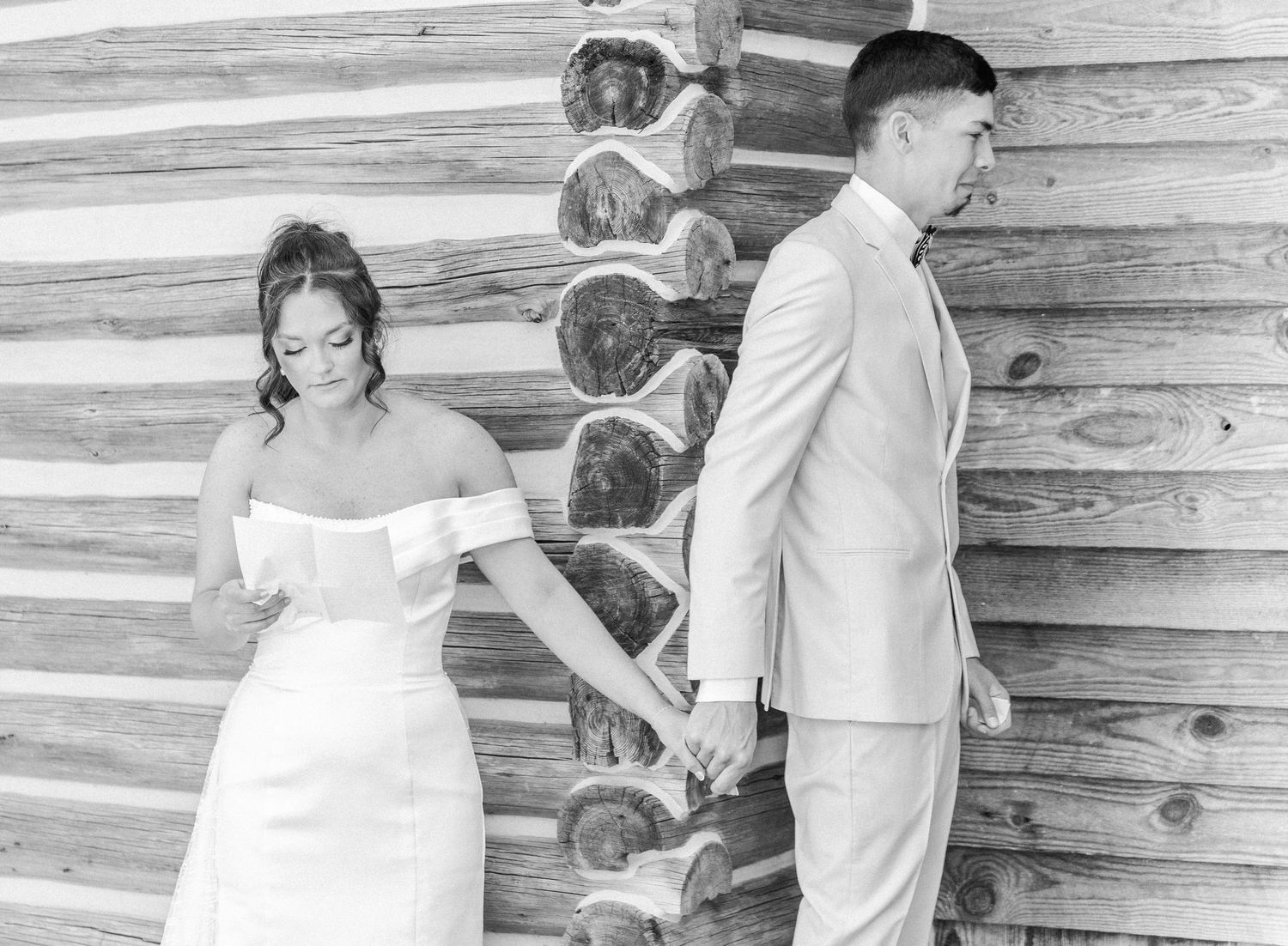 Black and white photo of bride and groom holding hands during an emotional first look by log wall.