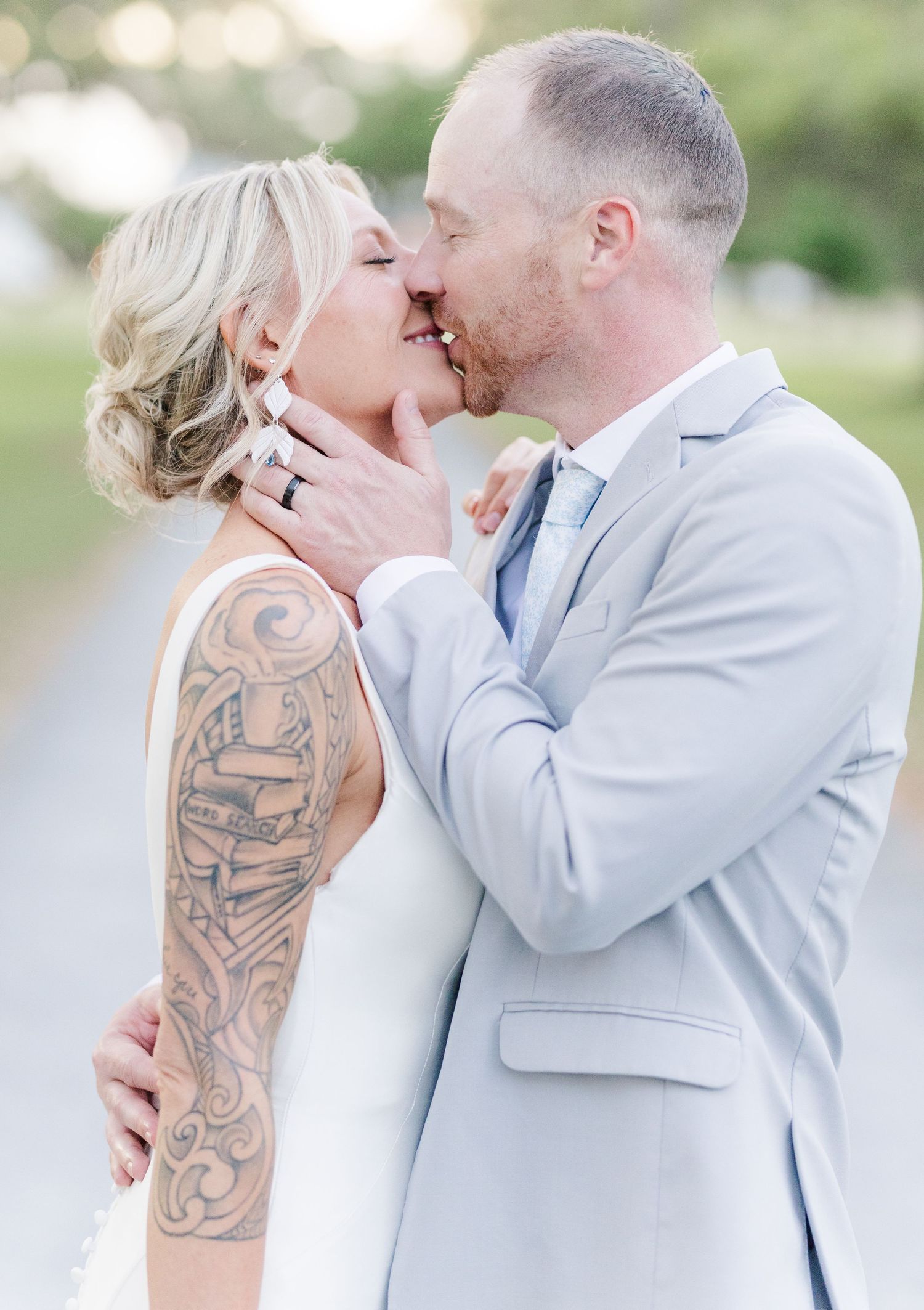 Bride with sleeve tattoo and groom in light gray suit share a tender kiss outdoors at their wedding.