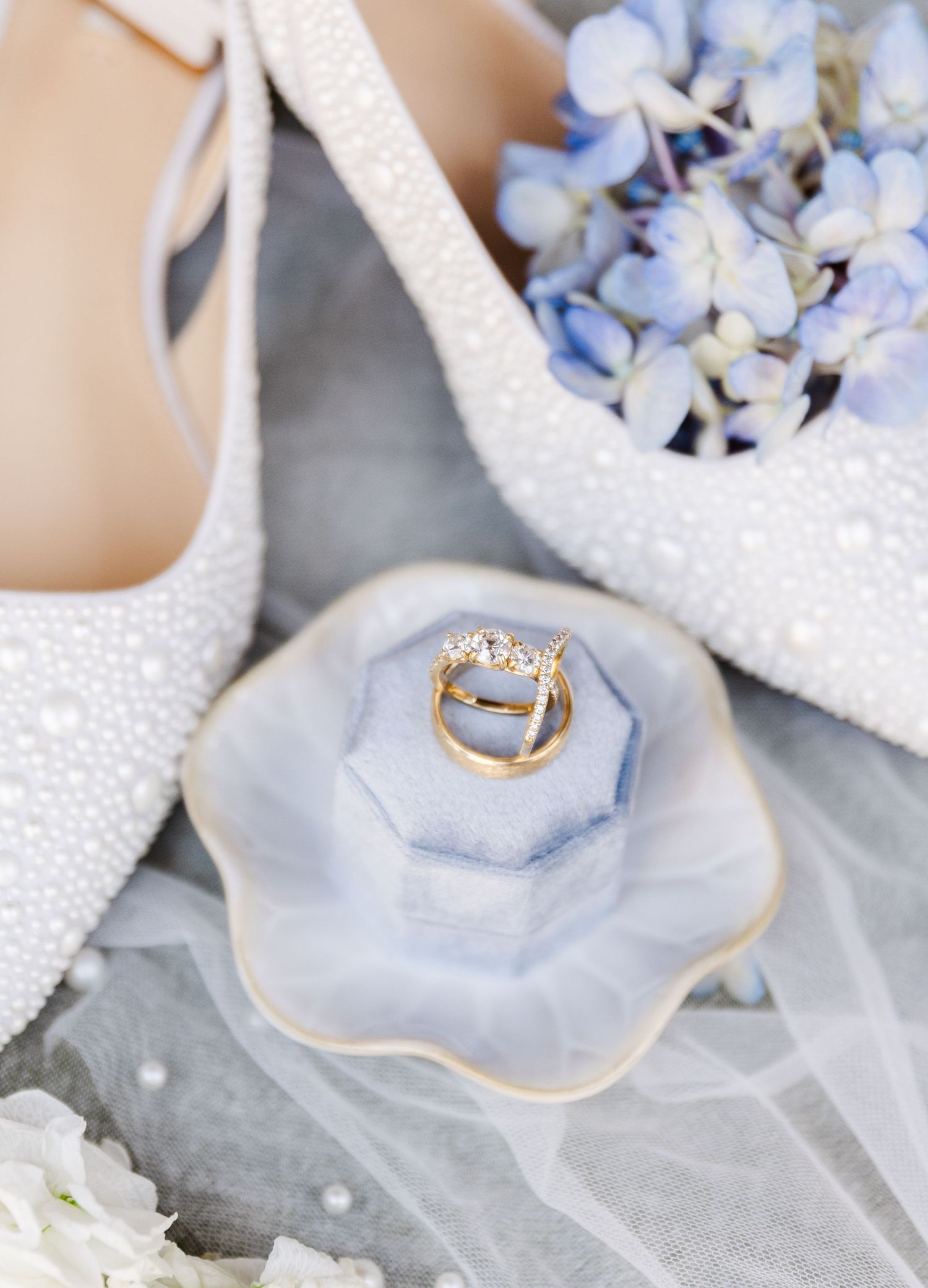 Pearl wedding heels beside blue hydrangeas and a floral dish holding gold diamond engagement and wedding bands.
