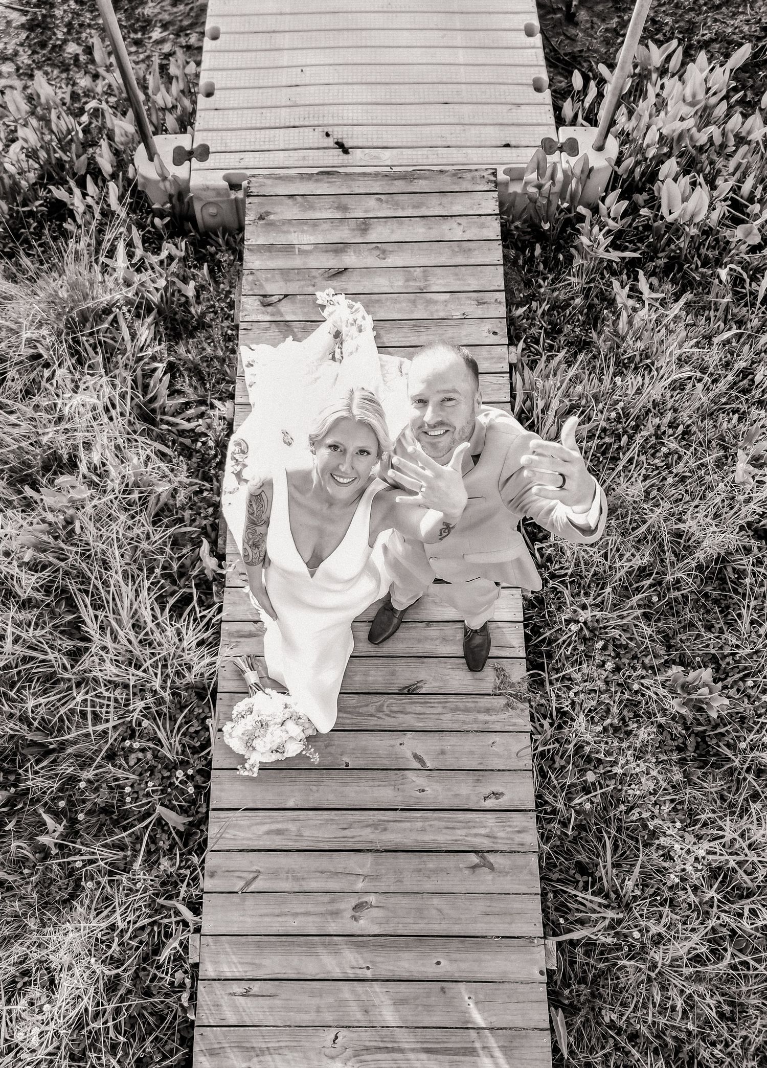 Aerial view of newlyweds waving on wooden boardwalk surrounded by greenery in black and white wedding photo.