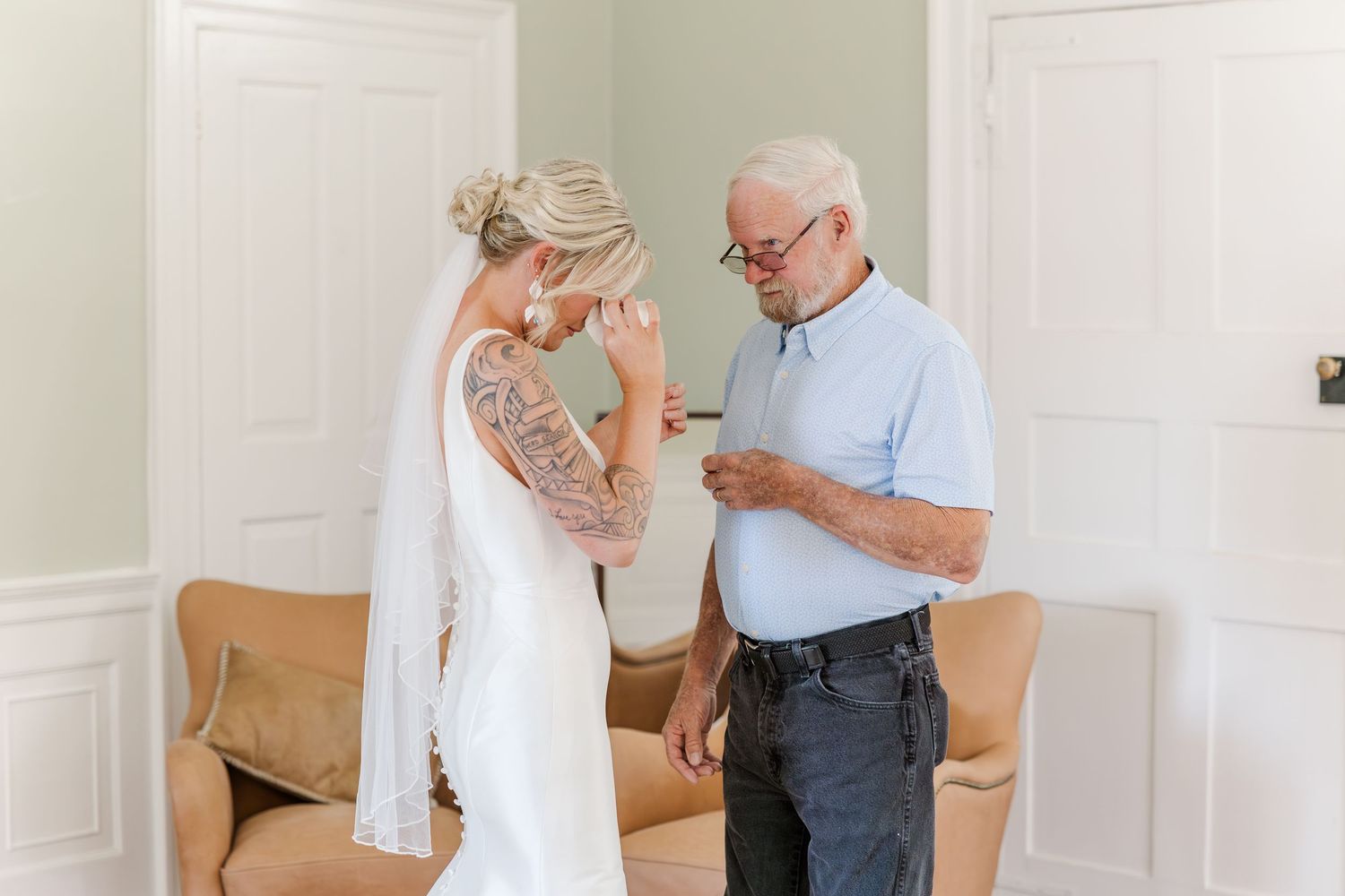 Bride in white wedding dress and veil shares emotional moment with elderly father figure indoors before ceremony.