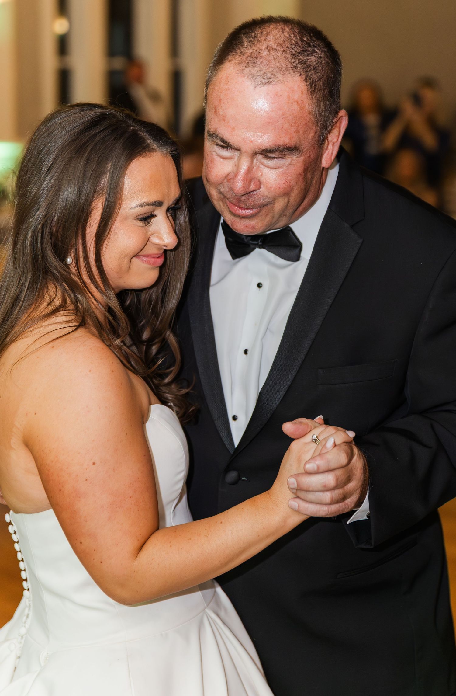 Bride and father share an emotional first dance at wedding reception, both smiling in formal attire.