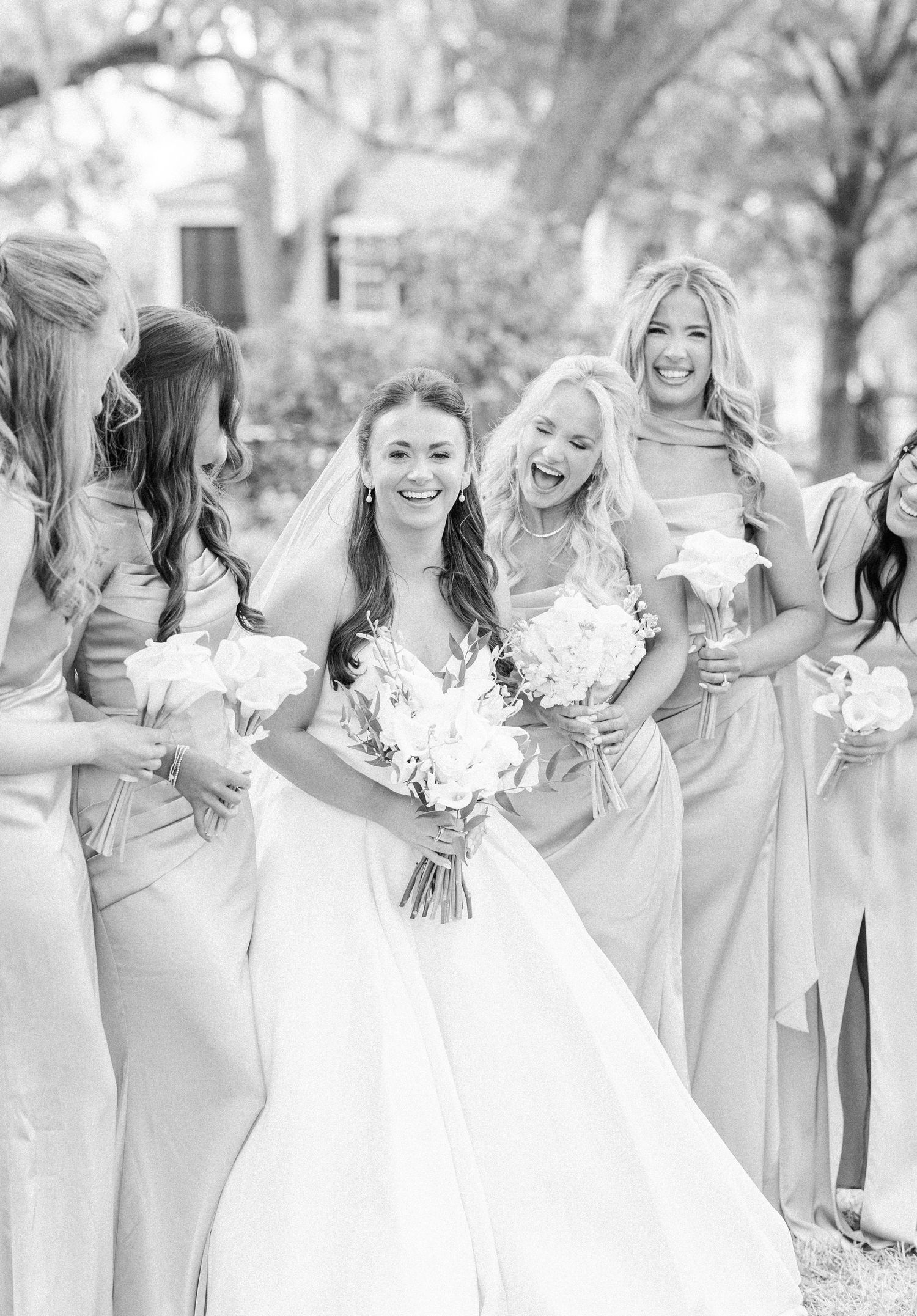 Black and white photo of laughing bride and bridesmaids in elegant gowns holding white floral bouquets.
