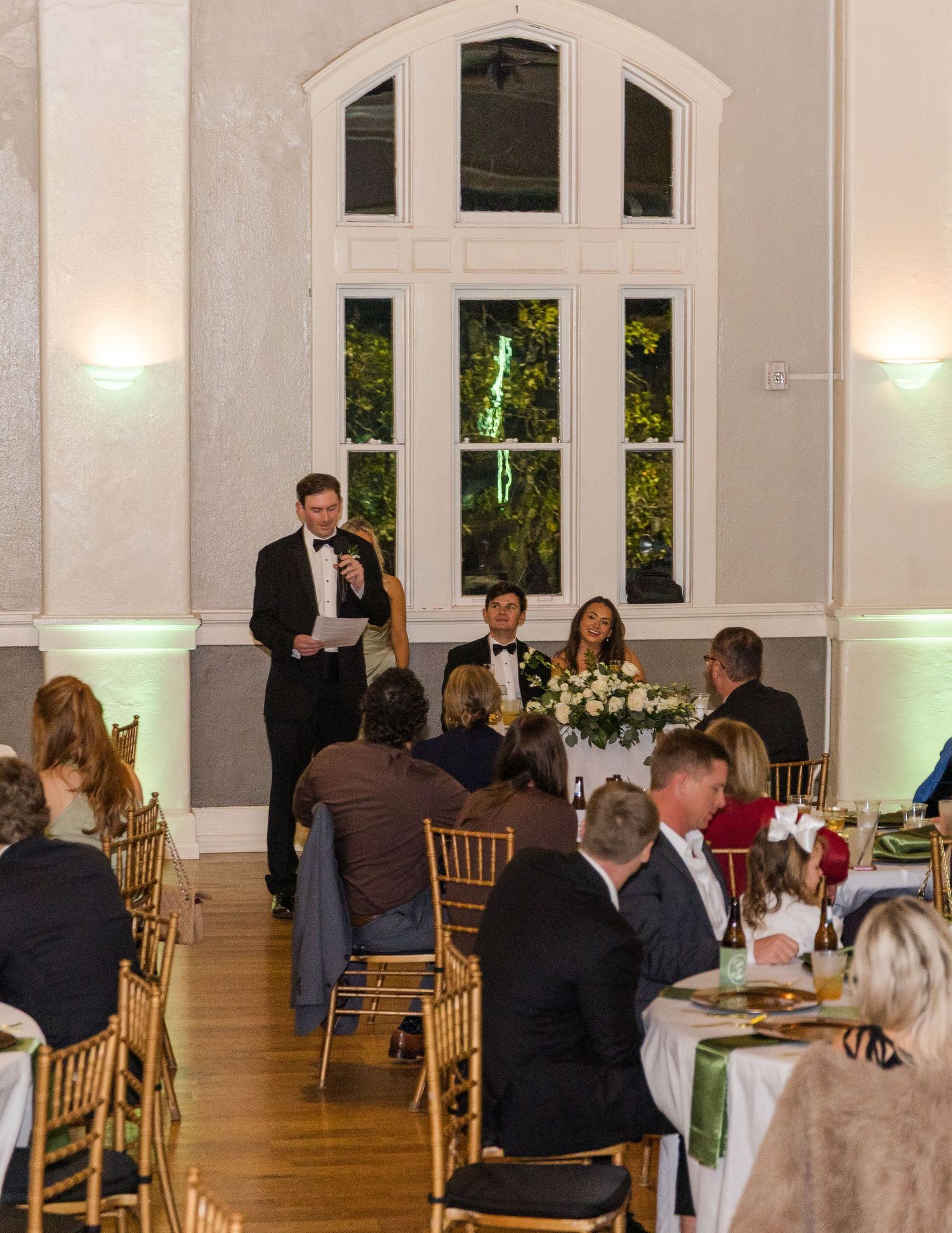 Elegant wedding reception in a grand hall with guests seated at tables while a man in a tuxedo gives a toast.