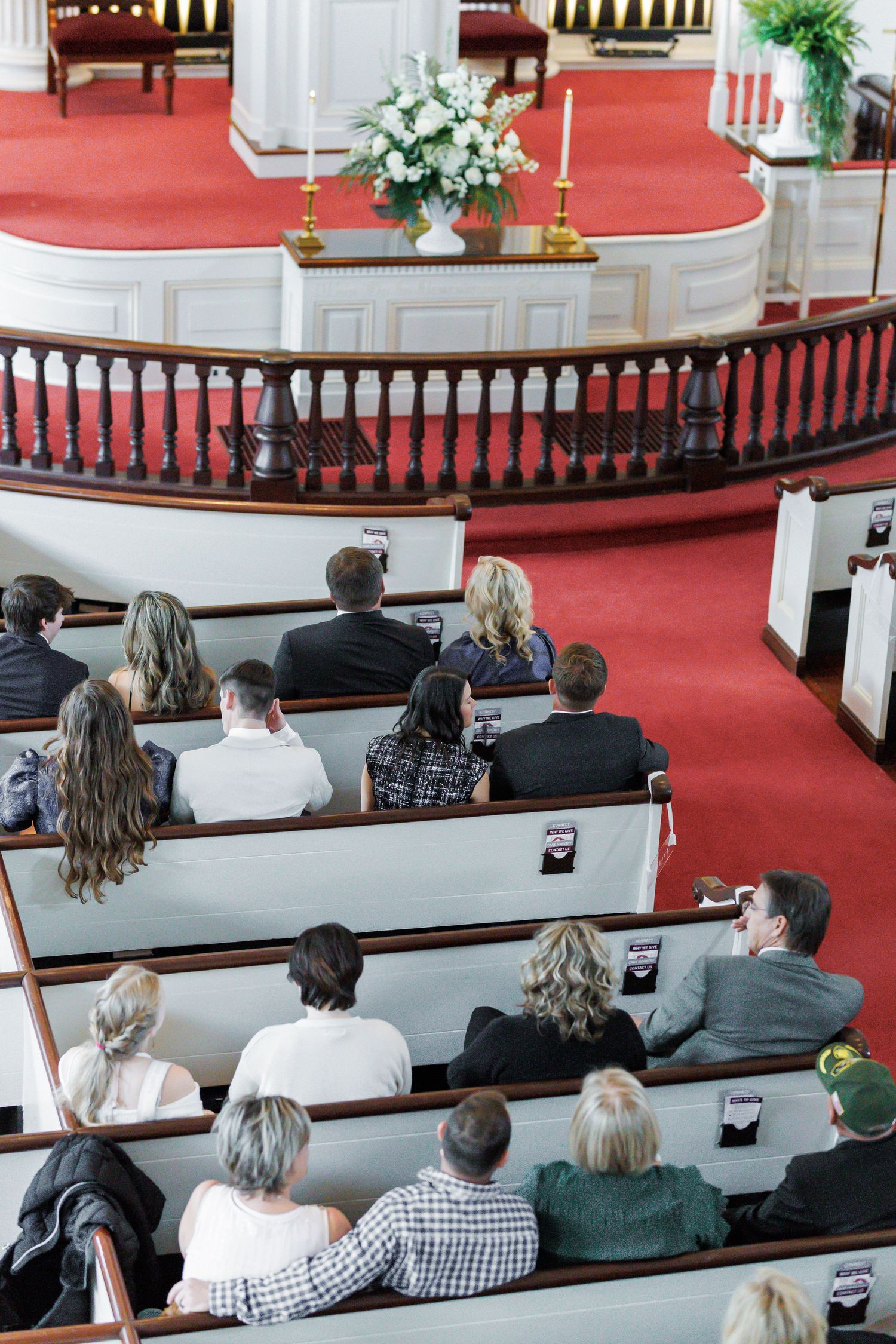 Guests seated in white wooden church pews during a wedding ceremony with red carpet altar.