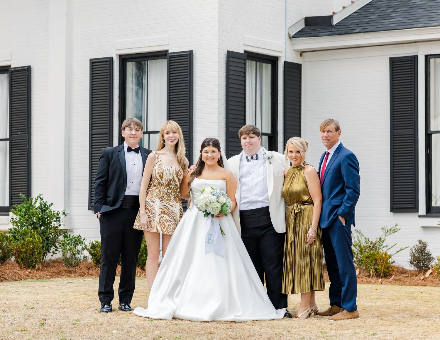 Wedding party of six posing outside white building with black shutters in formal attire.