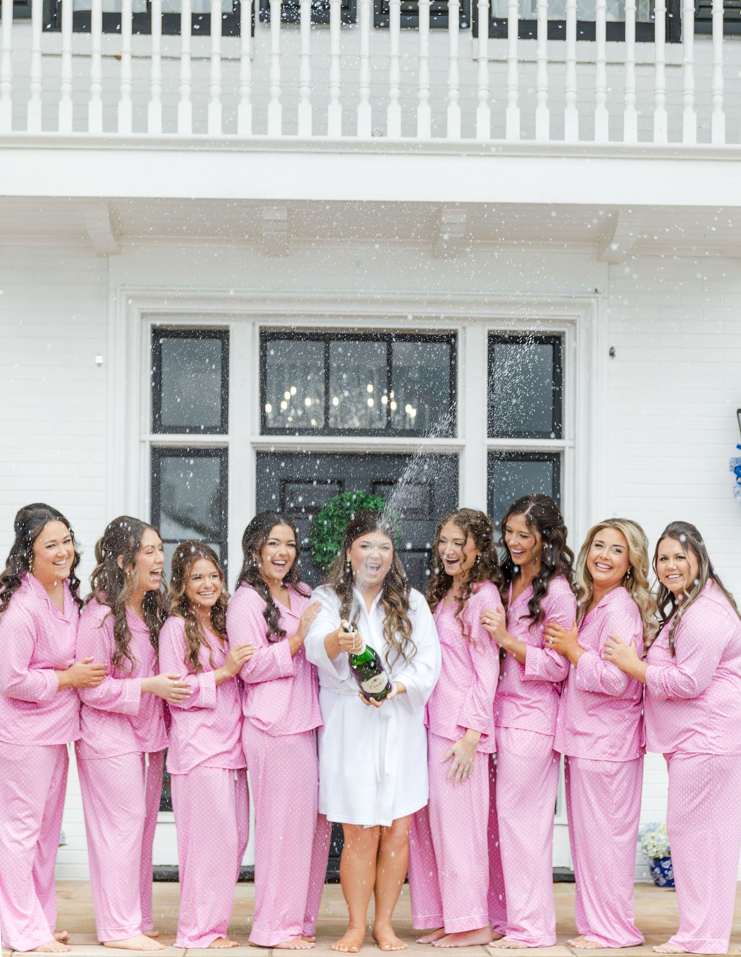 Bride in white robe pops champagne with bridesmaids in matching pink pajamas during wedding morning preparations.