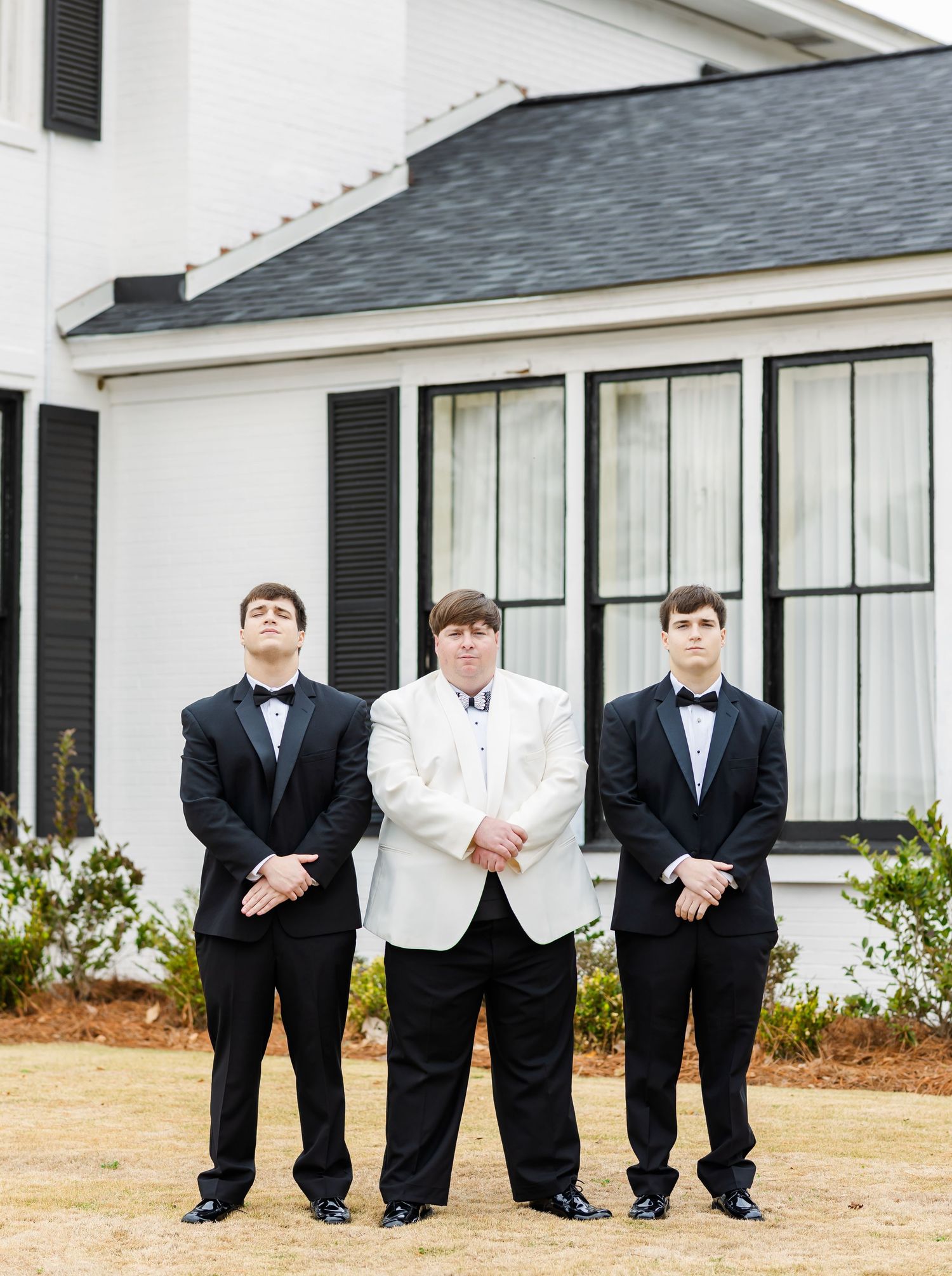 Groom in white jacket flanked by two groomsmen in black tuxedos standing outside a white wedding venue.