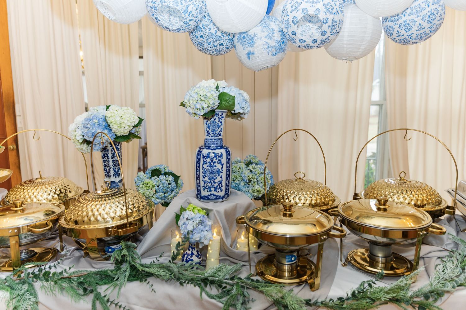 Gold chafing dishes on a wedding reception buffet table adorned with blue and white chinoiserie vases and hydrangea centerpieces.
