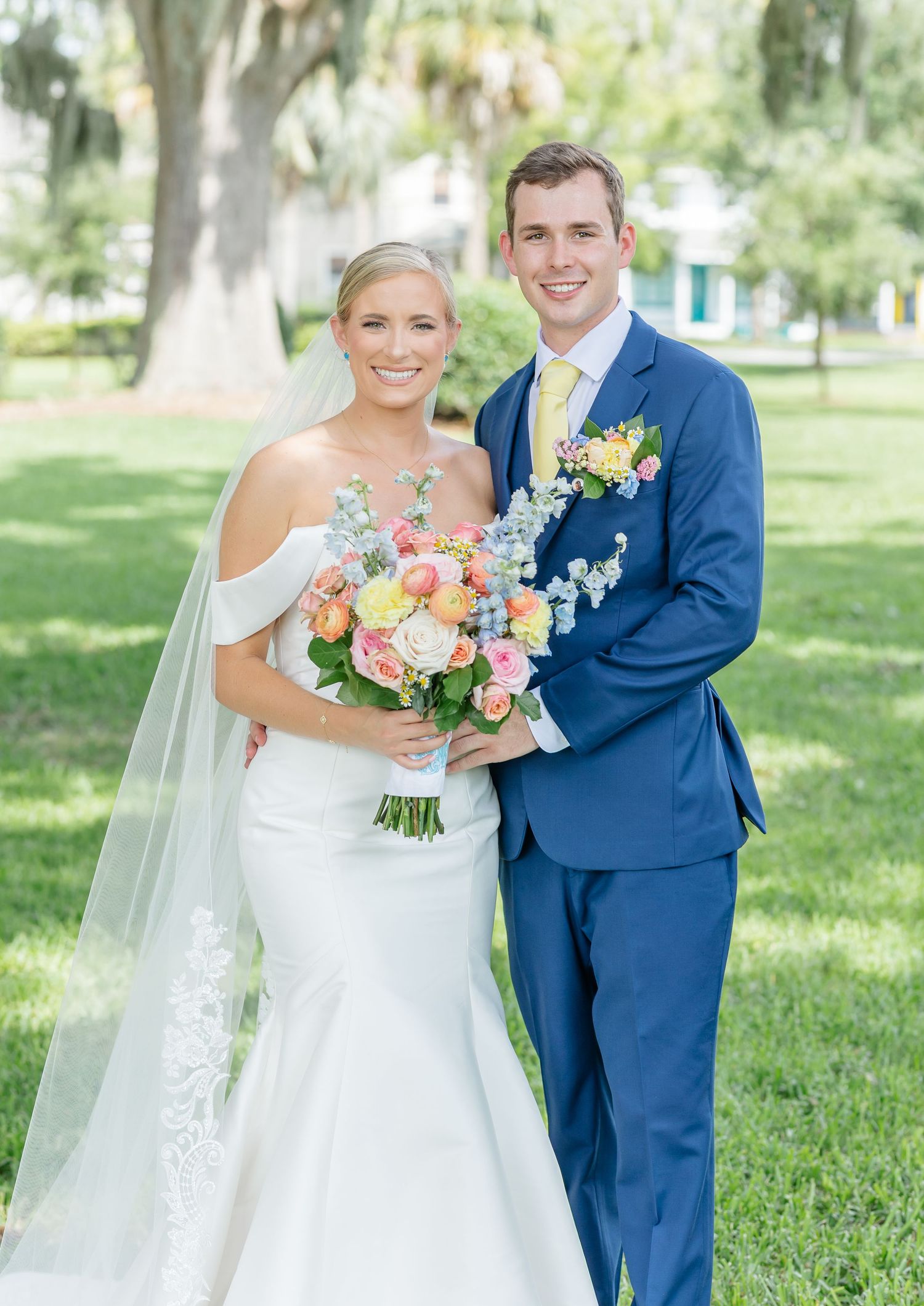 A wedding couple in formal attire pose outdoors with the bride holding a colorful bouquet against a sunny park backdrop.