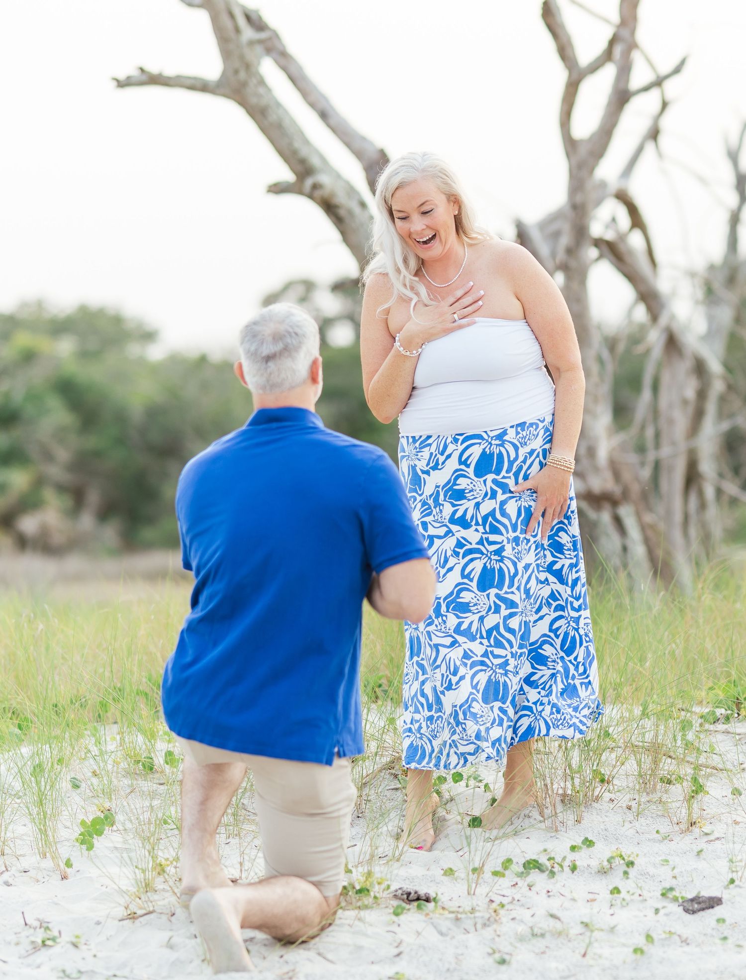A romantic proposal moment captured on a beach with white sand and bare trees, featuring a blue shirt and floral dress.