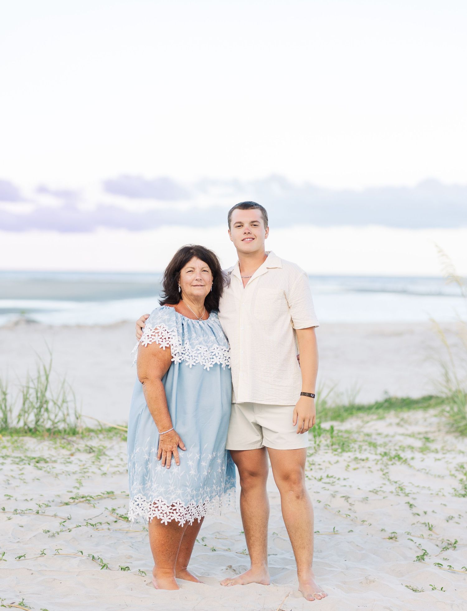 A couple stands barefoot on the beach wearing light summer attire during sunset.