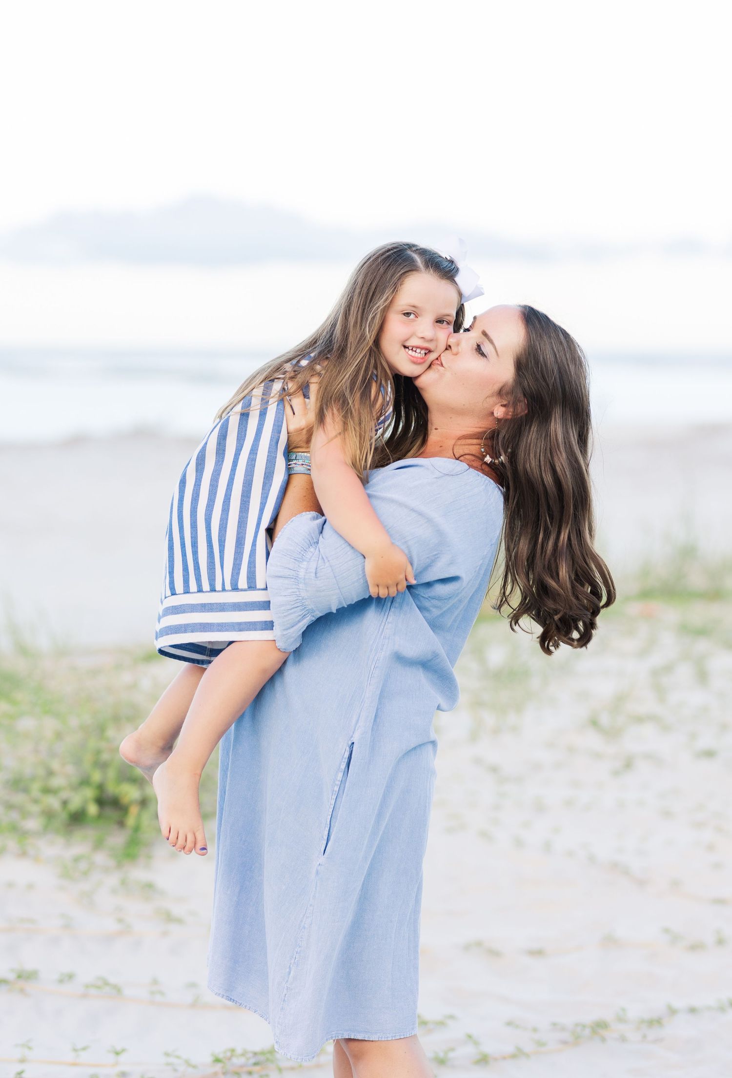 Mother and daughter share a joyful moment embracing on a misty beach while wearing blue coordinated outfits.