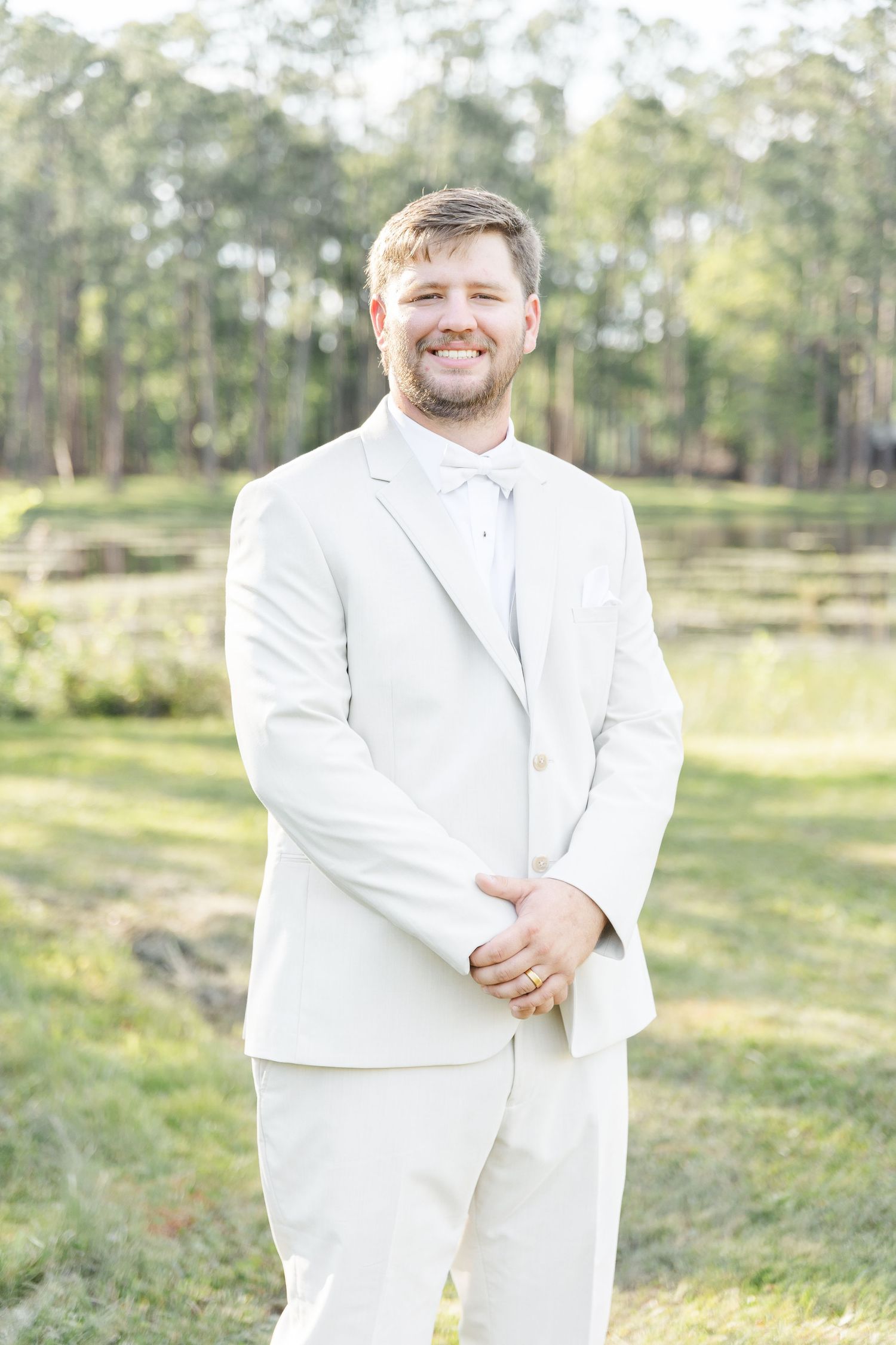Groom in white suit and bowtie smiles while standing in outdoor setting with trees and pond in background.
