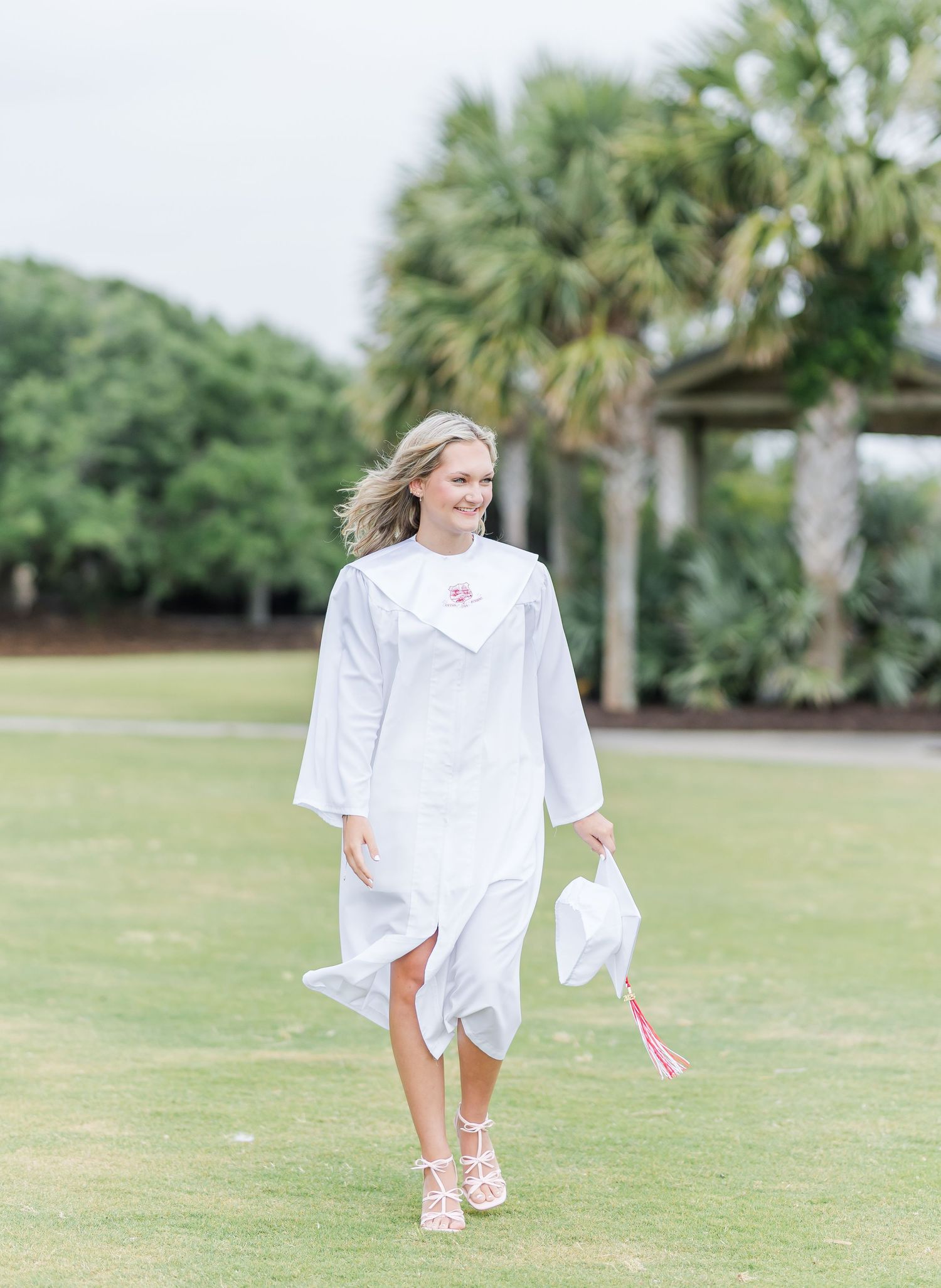 A graduate walks on grass holding a cap while wearing strappy sandals and white regalia.