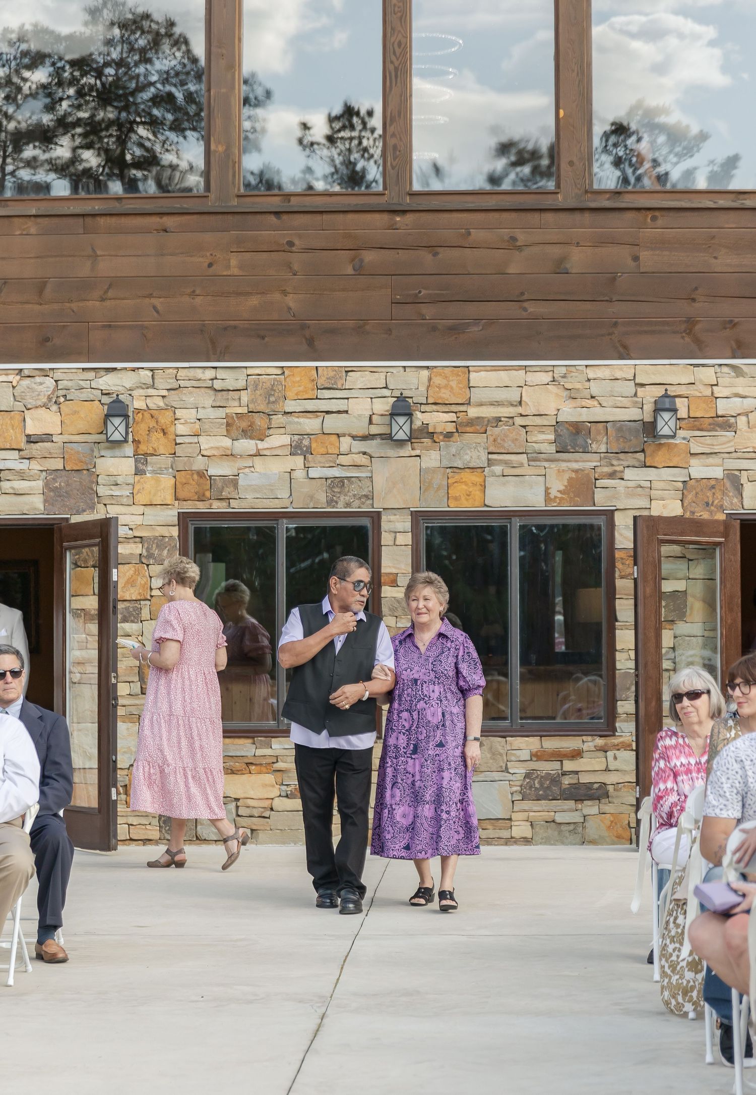 Wedding ceremony procession at outdoor stone venue with seated guests in white chairs.