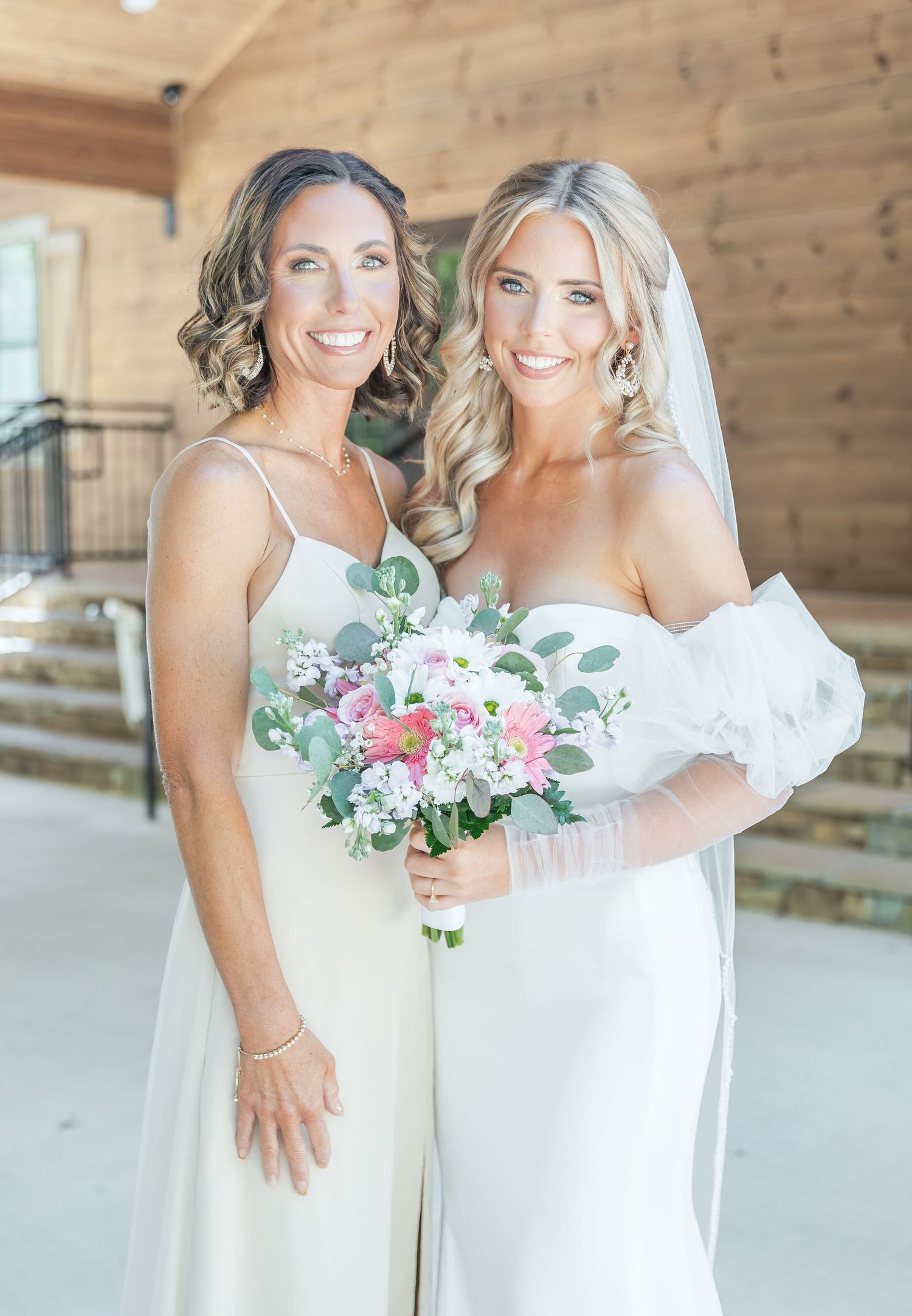 Two women in white dresses share joyful moment at rustic wedding venue.