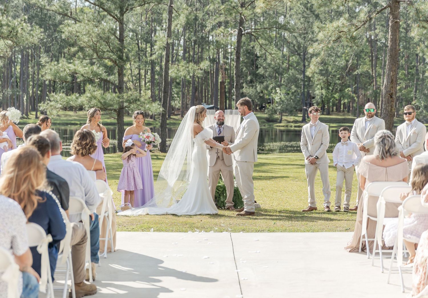 Wedding ceremony takes place on sunny day with pine forest backdrop and seated guests.