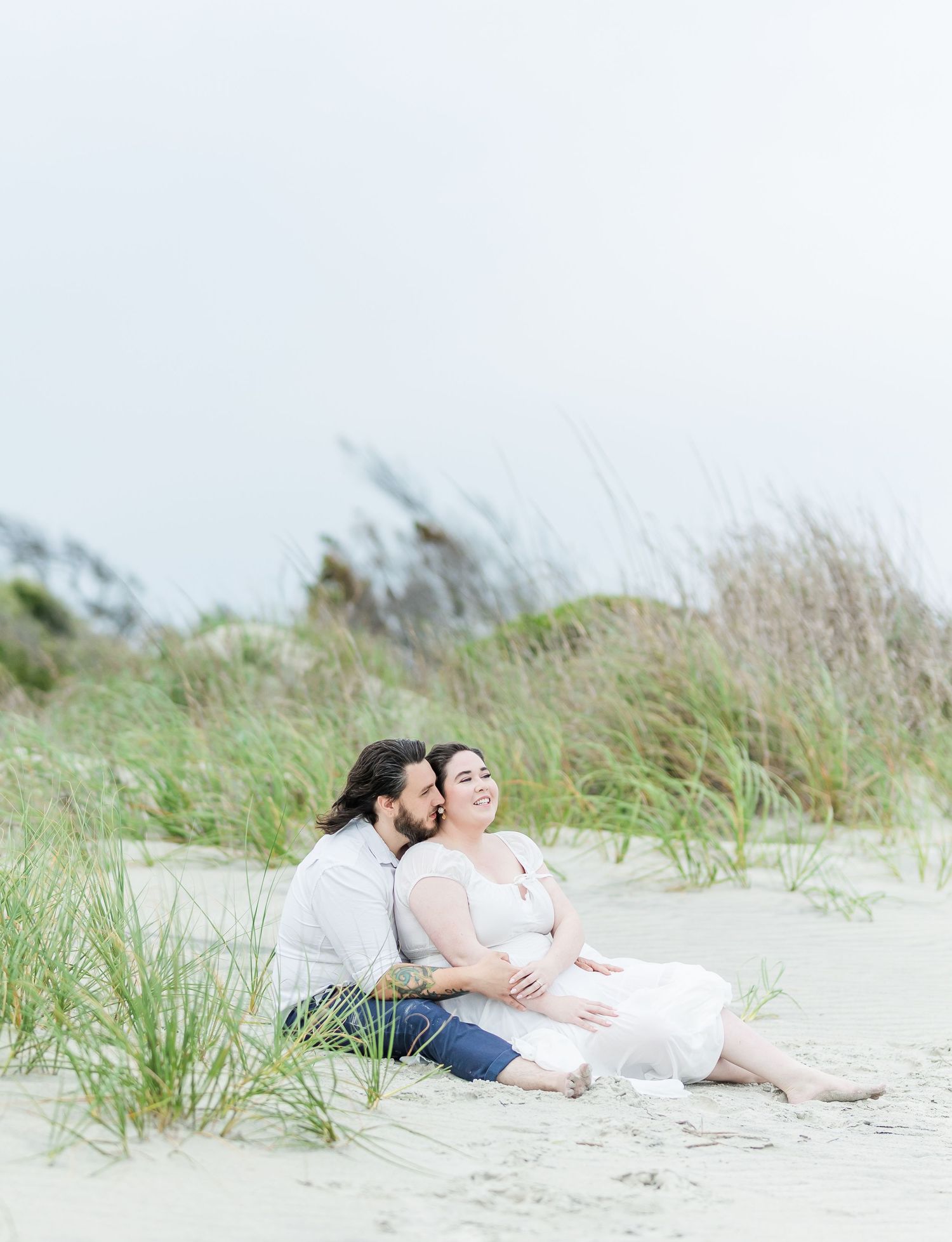 A romantic couple sits together on a sandy beach with tall grass swaying in the breeze.