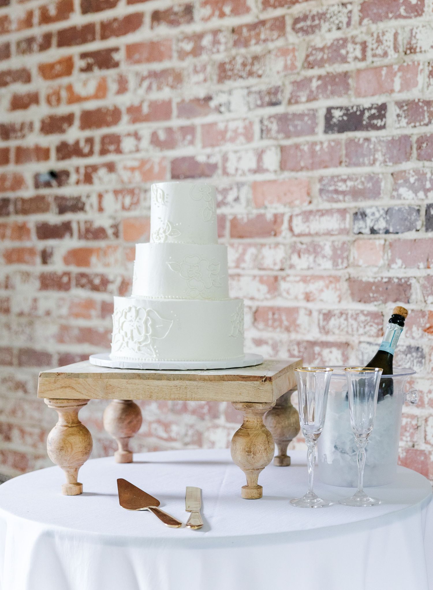 Elegant four-tier white wedding cake on wooden stand against rustic brick wall.