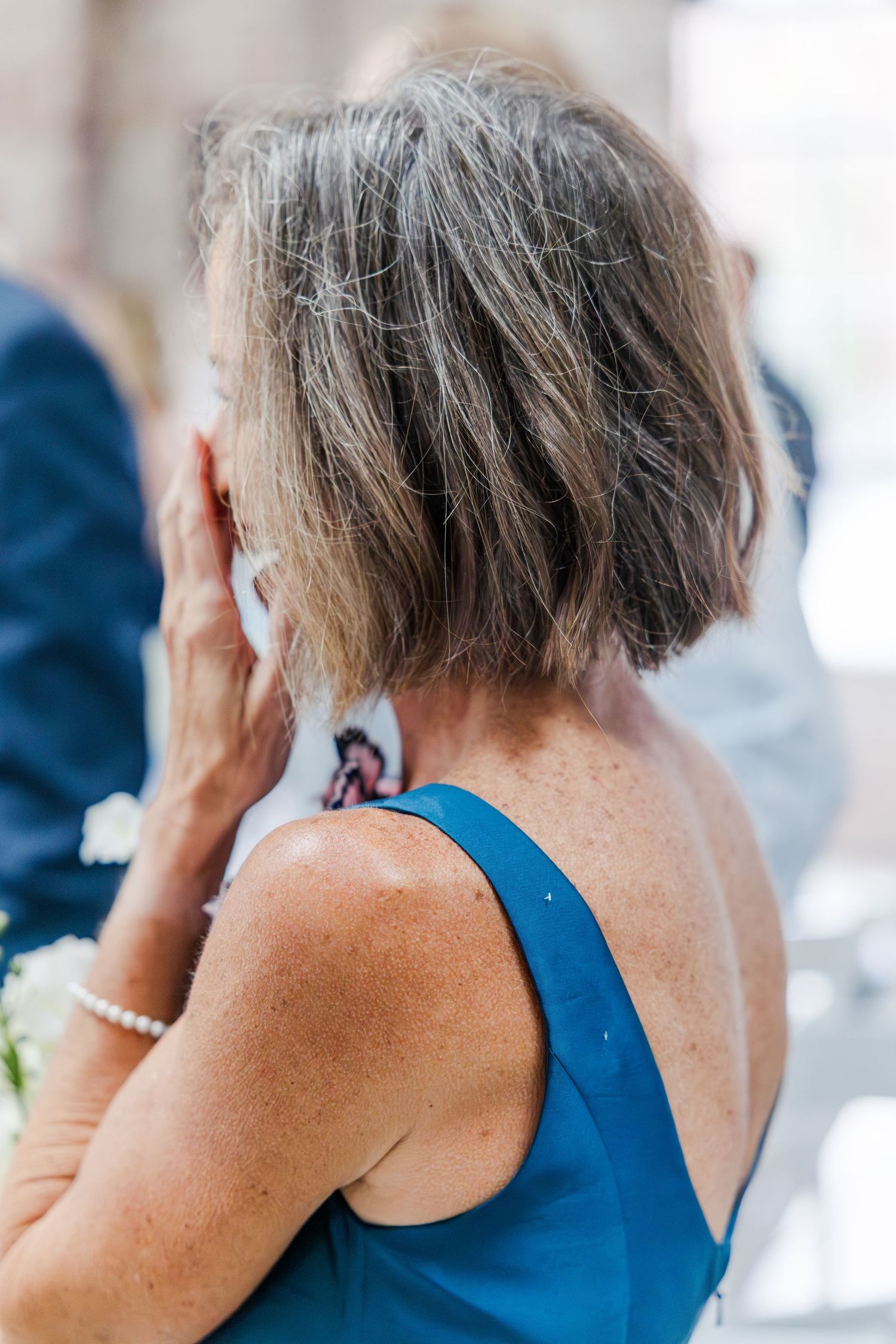 Woman in teal dress with gray bob hairstyle wiping tear at wedding ceremony.