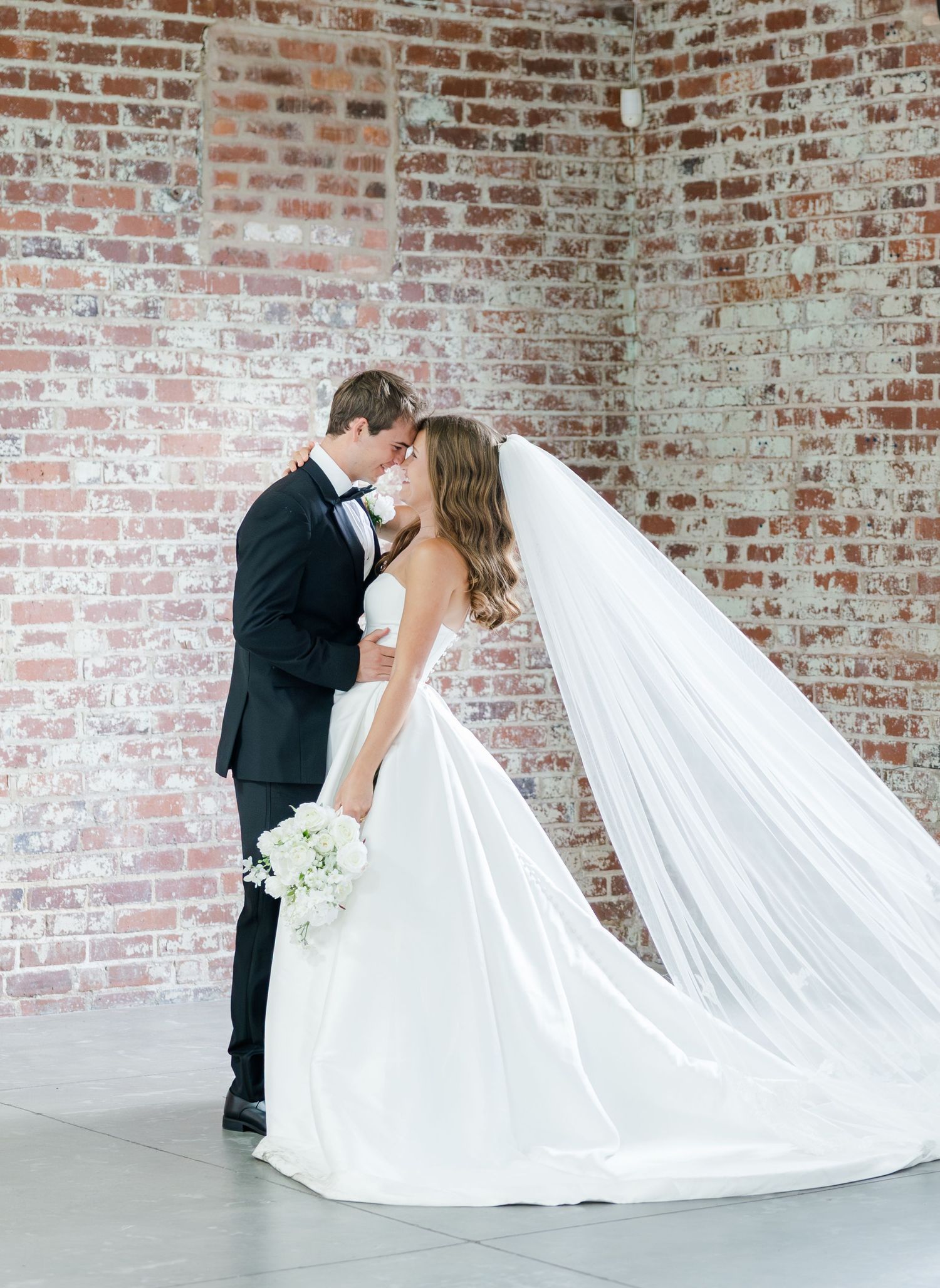 Bride and groom kissing in front of brick wall, brides veil flowing dramatically to the side.