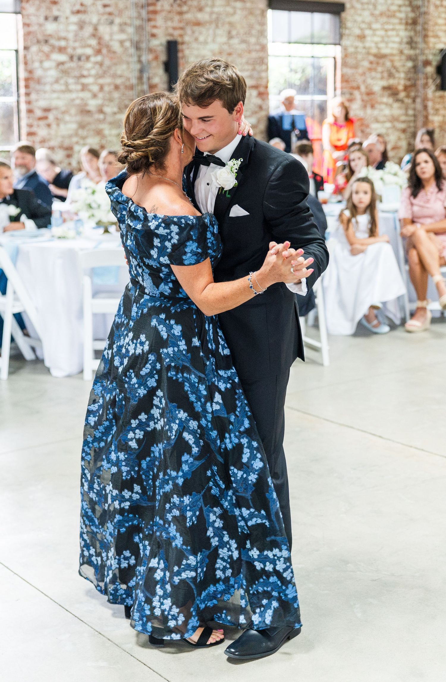Groom in black tuxedo shares mother-son dance with woman in blue floral gown.