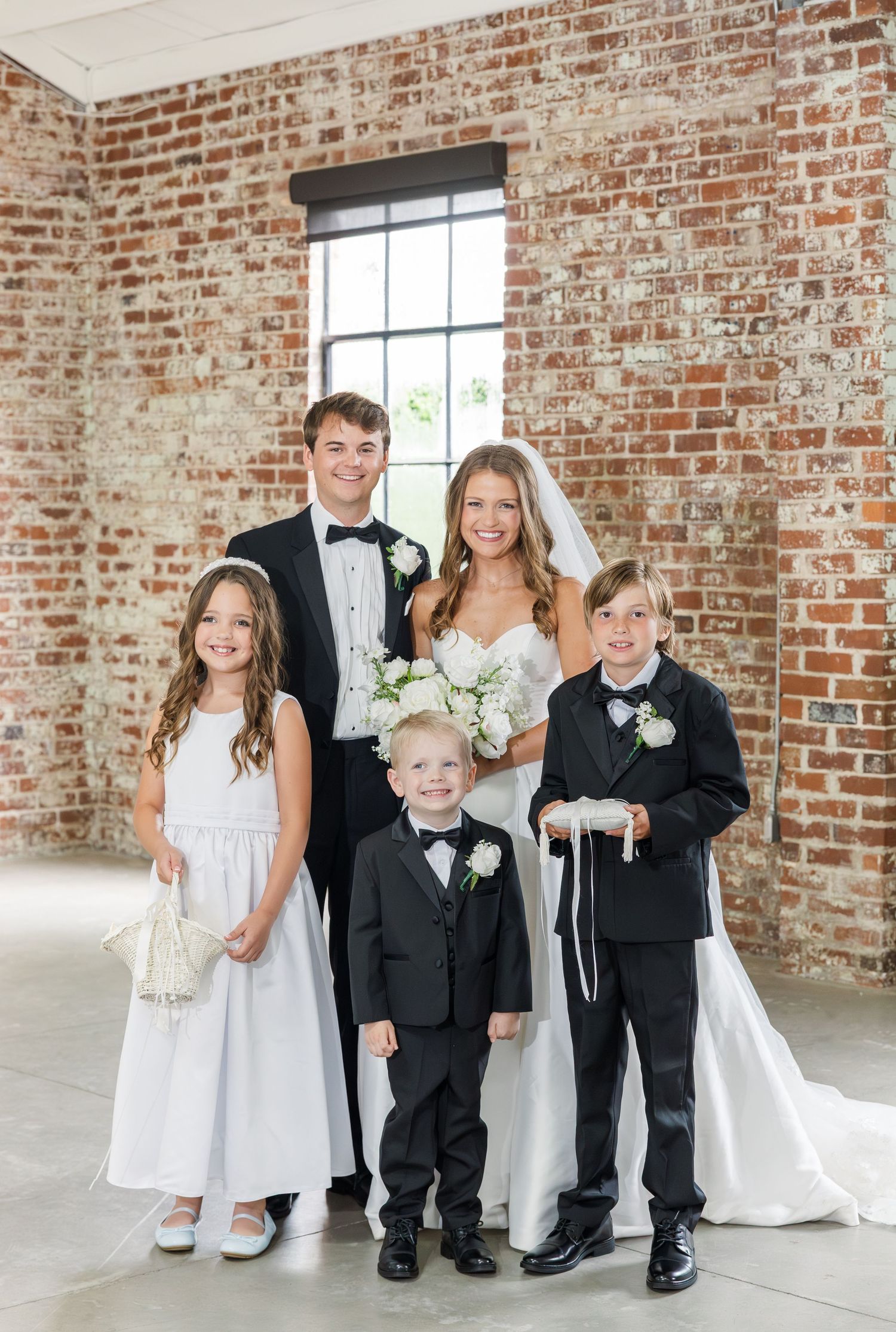 Bride and groom pose with flower girl, ring bearer, and young boy in matching black tuxedos.