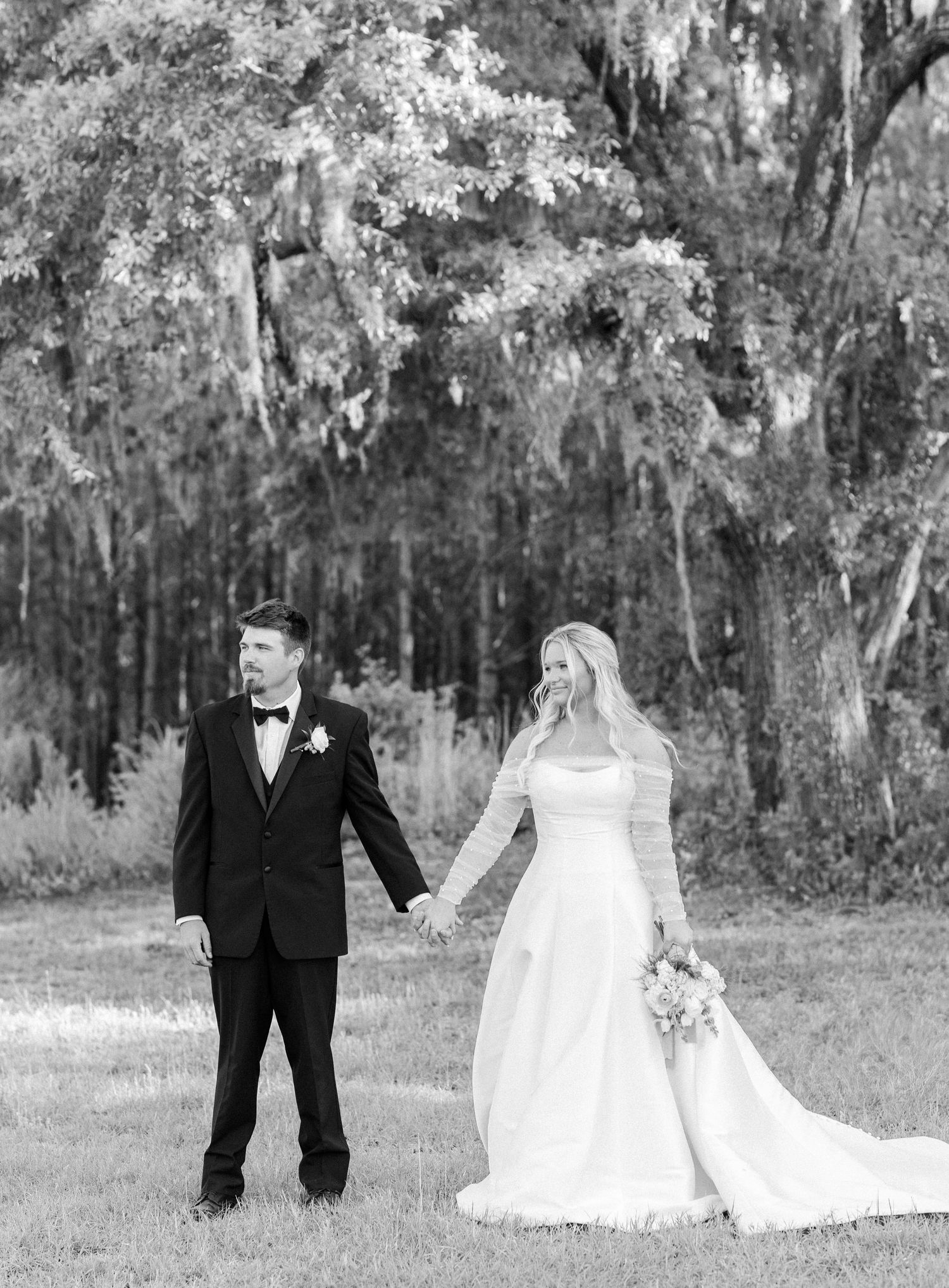 Bride and groom holding hands in a field with Spanish moss trees in a classic black and white wedding photo.