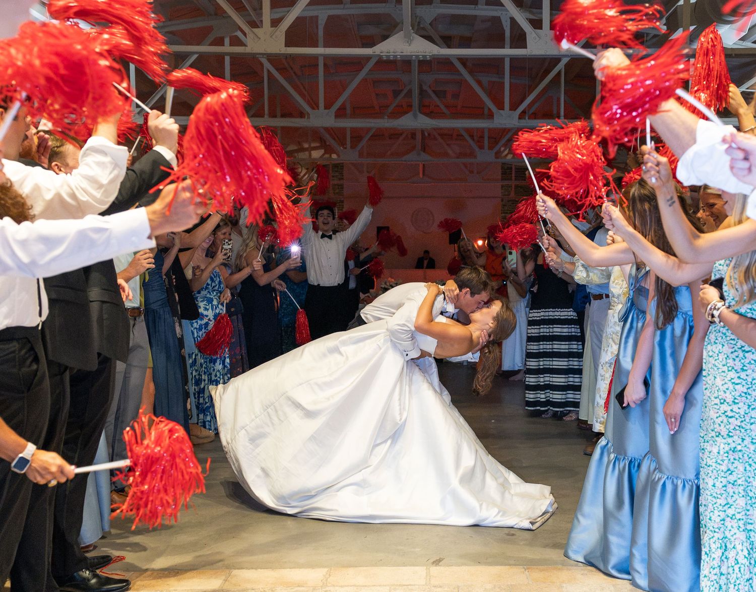Newlyweds share romantic dip kiss during grand exit with guests waving red feather fans at wedding celebration.