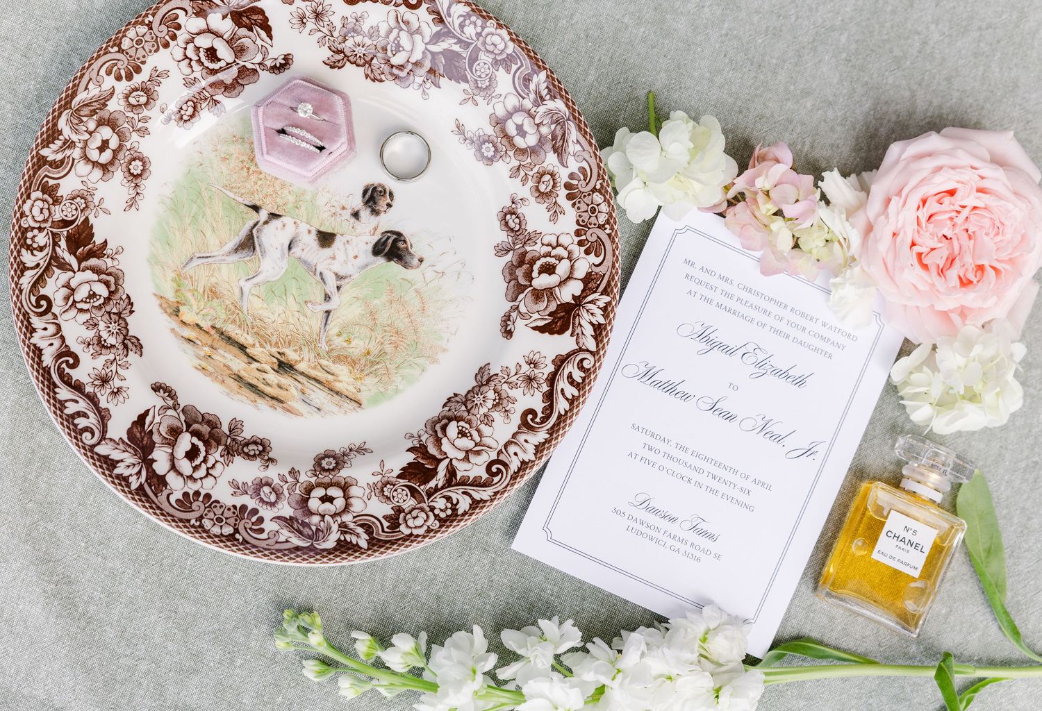 Elegant wedding flat lay with floral china plate, rings, invitation, pink roses, and Chanel perfume on gray background.