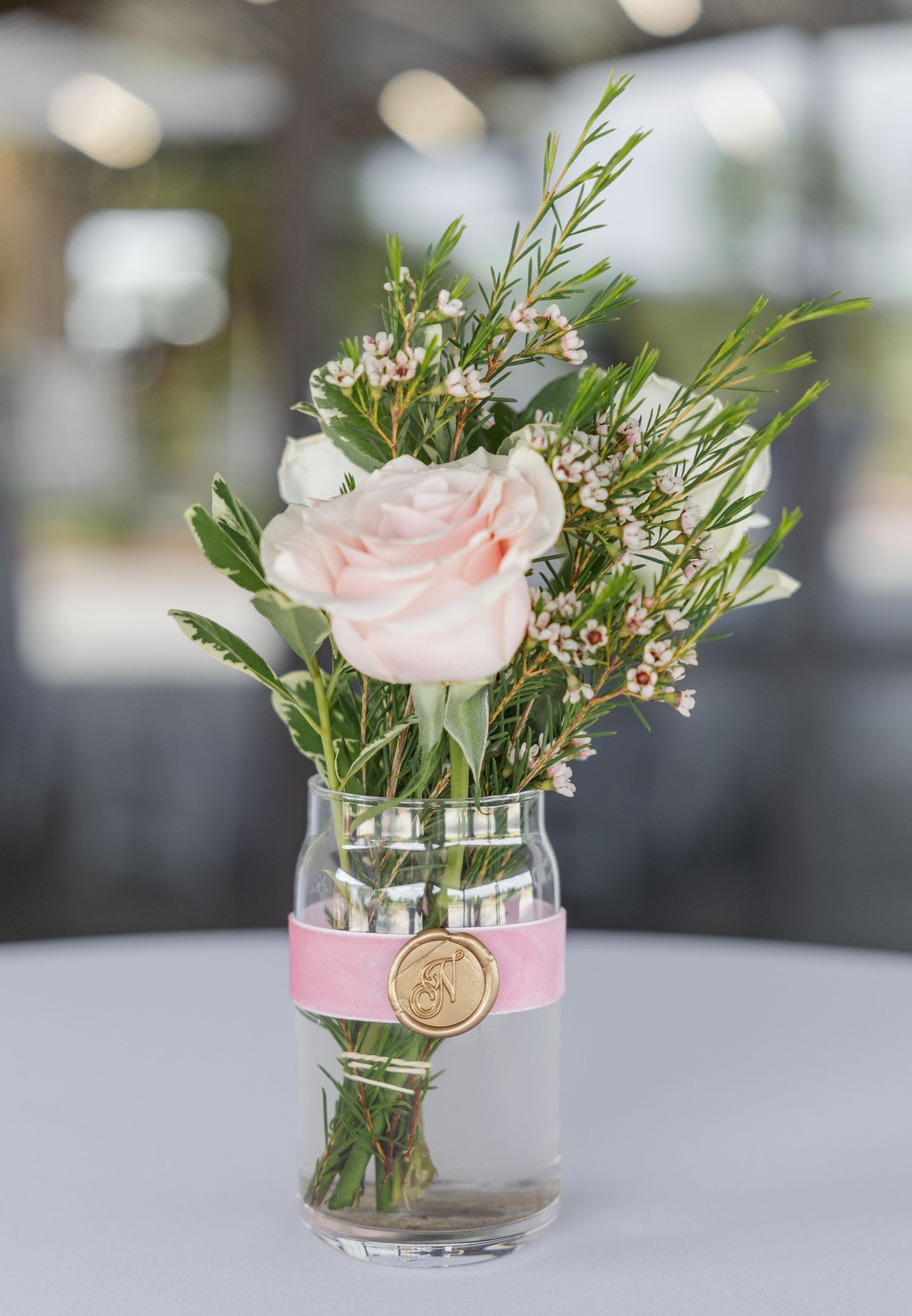 Delicate blush pink rose and wax flower arrangement in glass vase with pink ribbon and gold monogram seal.
