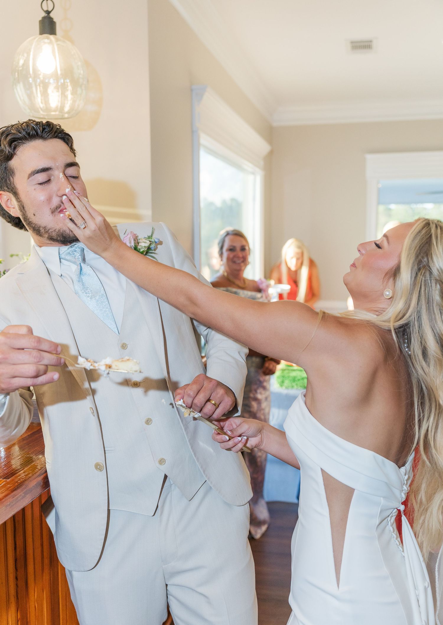 Bride playfully smashes cake into grooms face during wedding reception indoors.