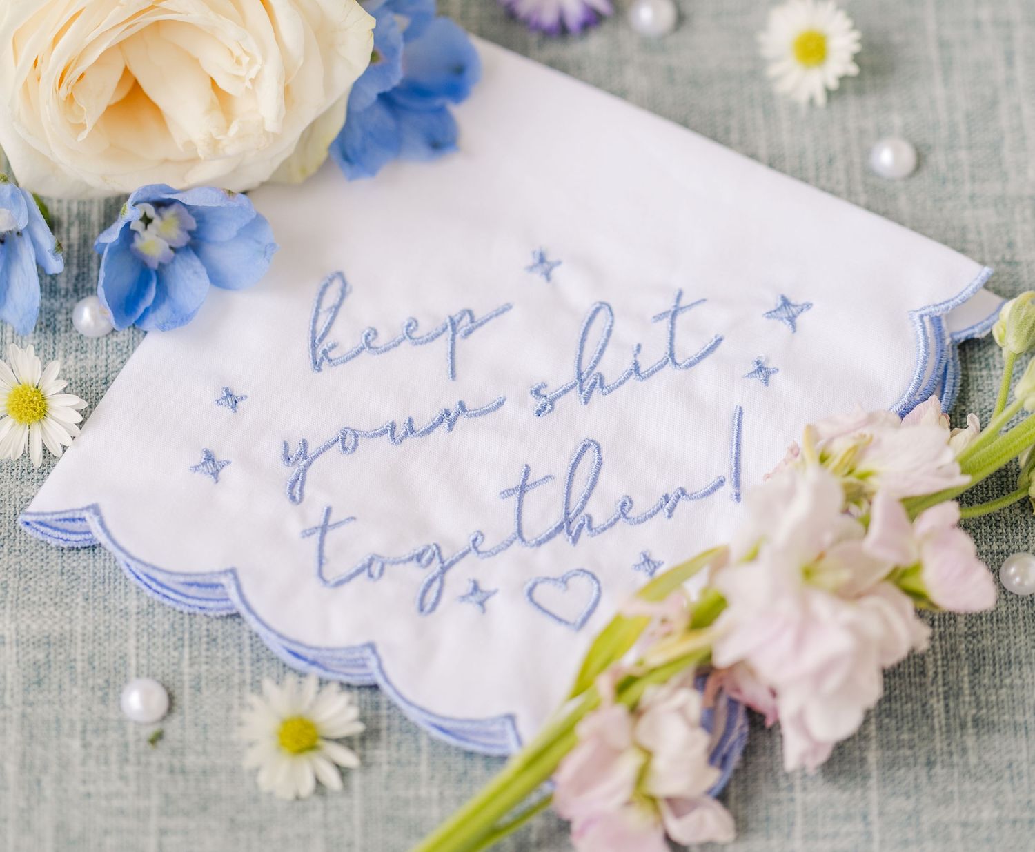 White scalloped handkerchief embroidered in blue with keep your shit together surrounded by flowers and pearls.