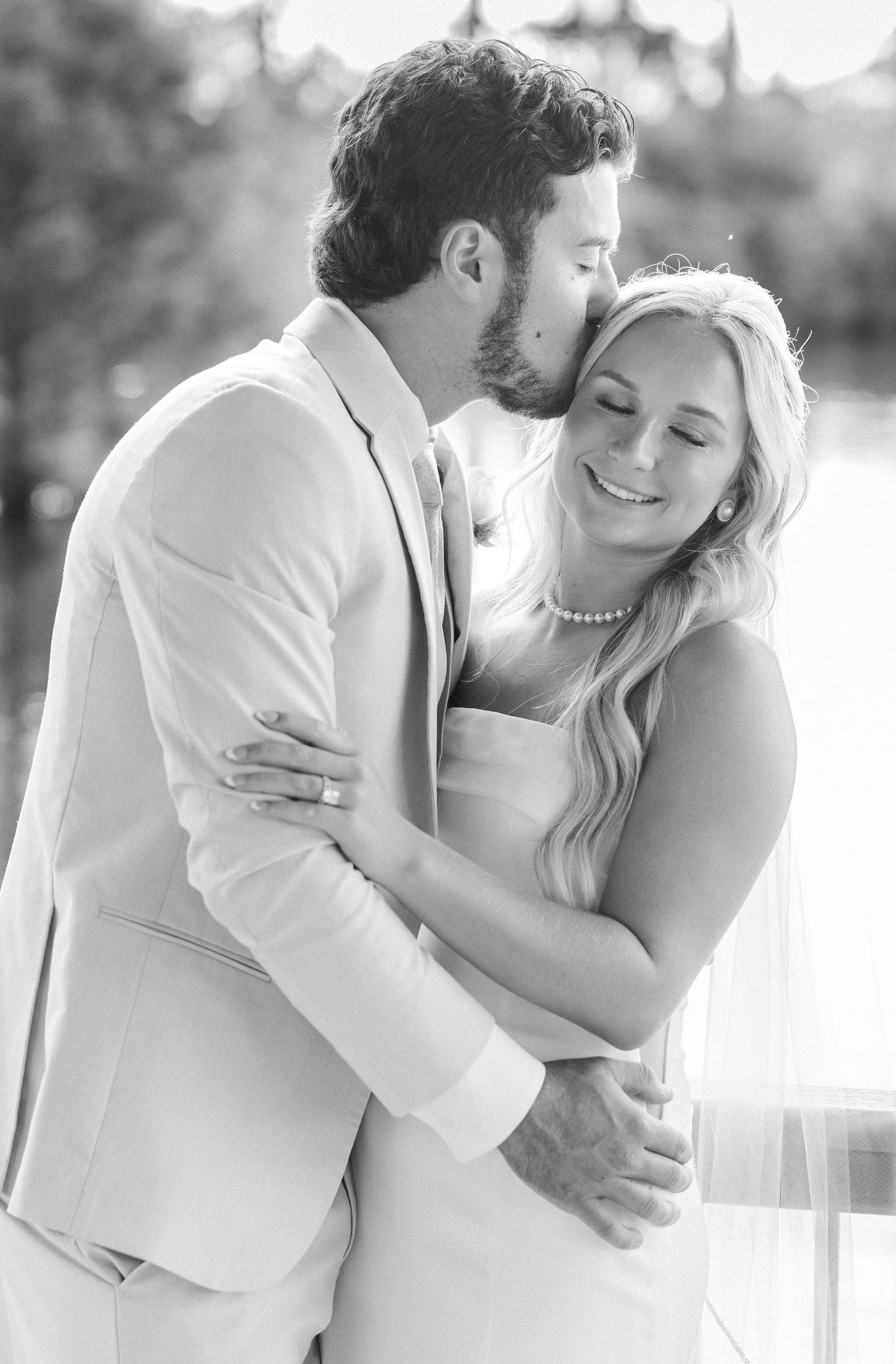 Black and white romantic portrait of groom kissing smiling bride on cheek in outdoor setting.